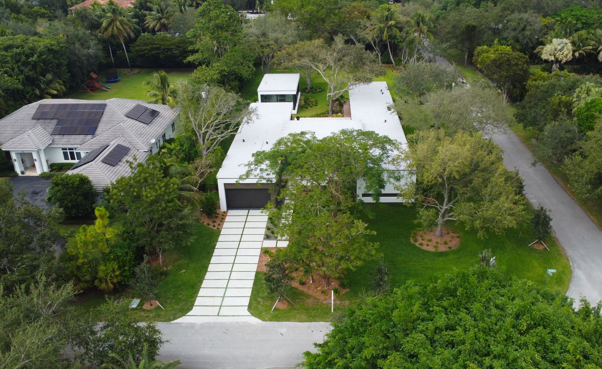 Aerial view of a modern white house with a flat roof, driveway, and surrounding green trees and foliage.