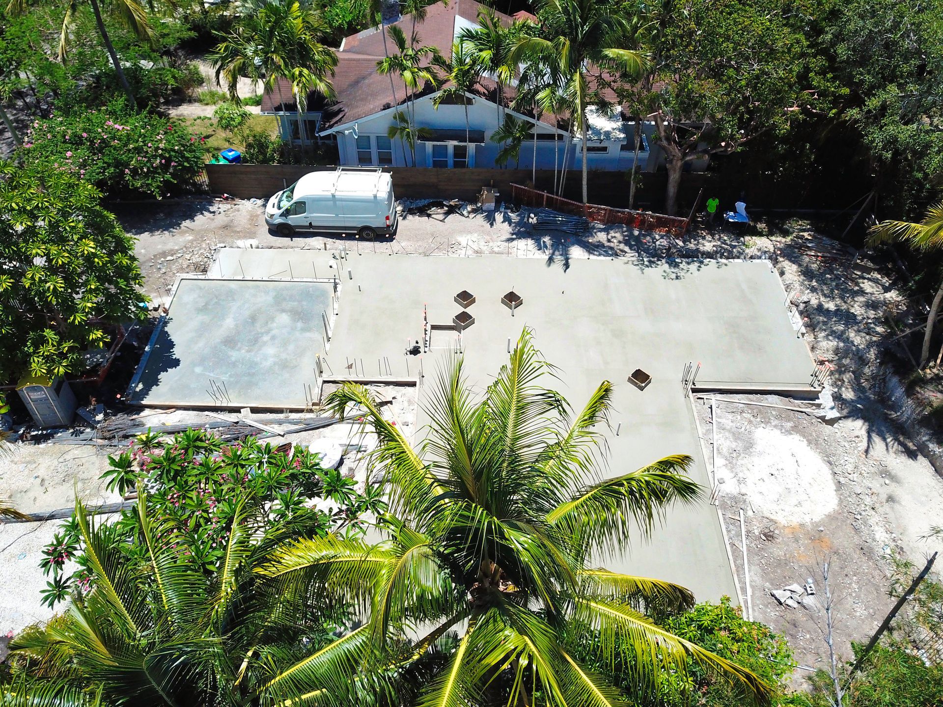 Aerial view of a concrete foundation with a white van, near a house with a brown roof and green foliage.