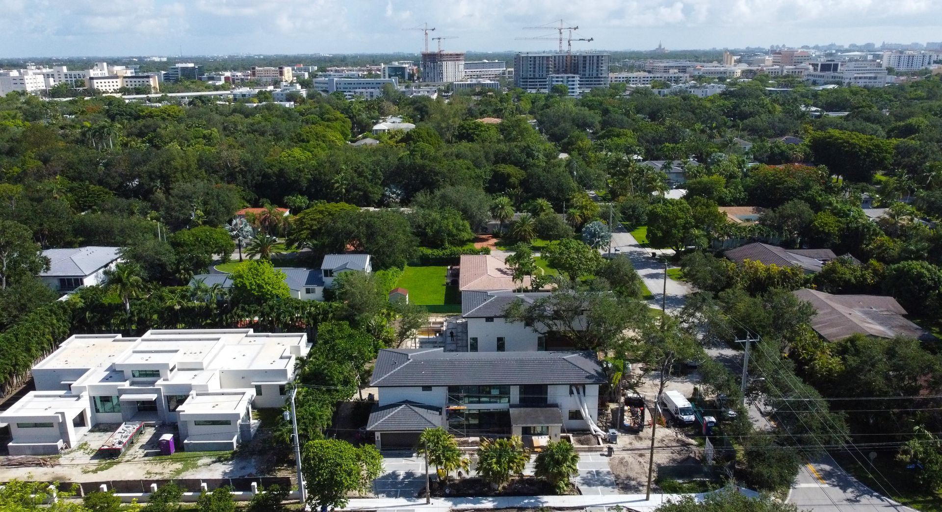 Aerial view of a suburban neighborhood with modern homes, lush trees, and city skyline in the background.