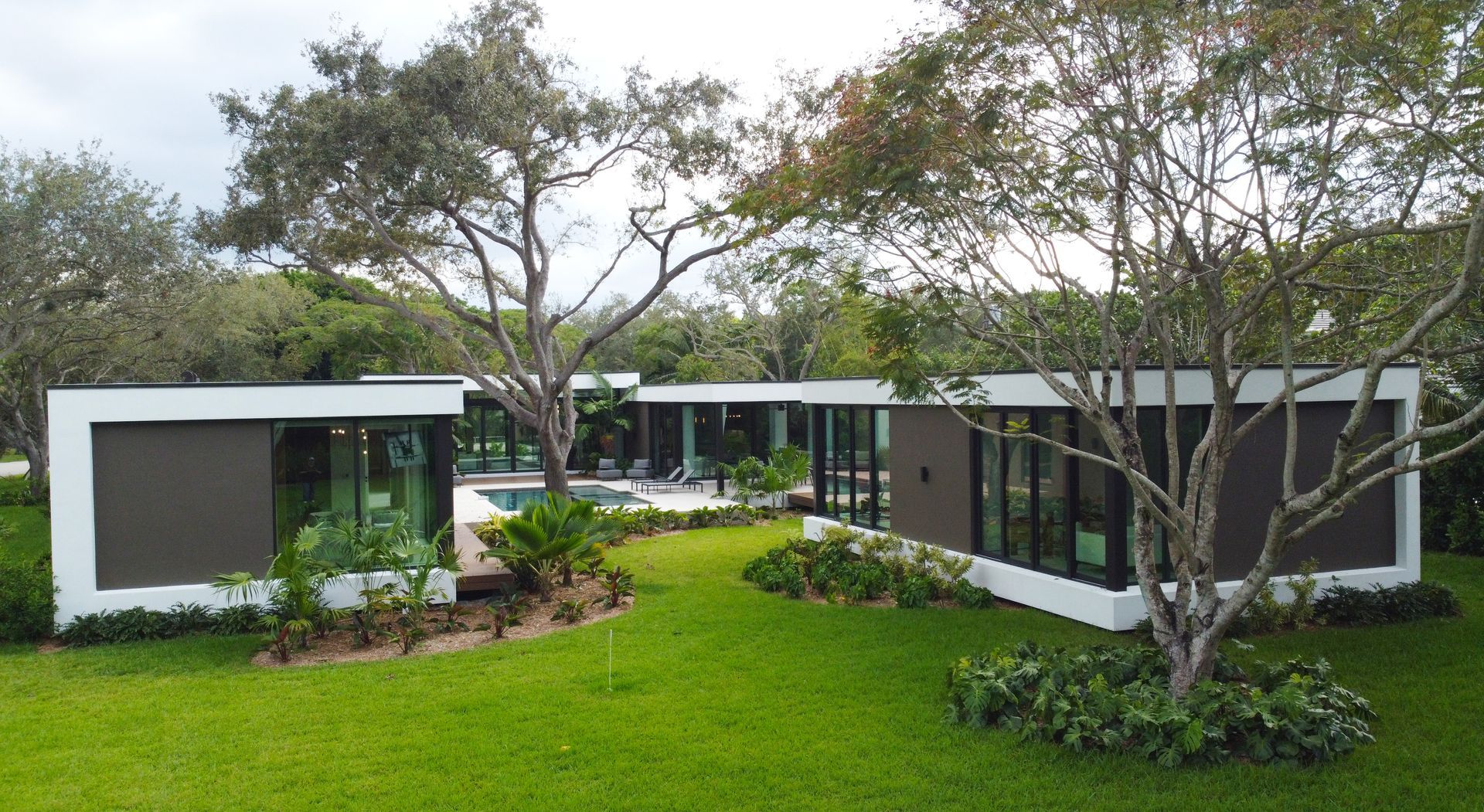 Modern, low-profile house with a flat roof and dark rectangular window frames, surrounded by green lawn and trees.