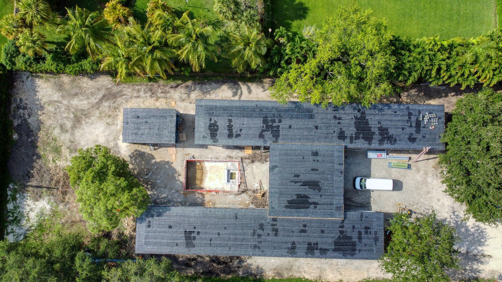 Overhead view of a house under construction with a black roof, surrounded by green trees and grass, with a white van.