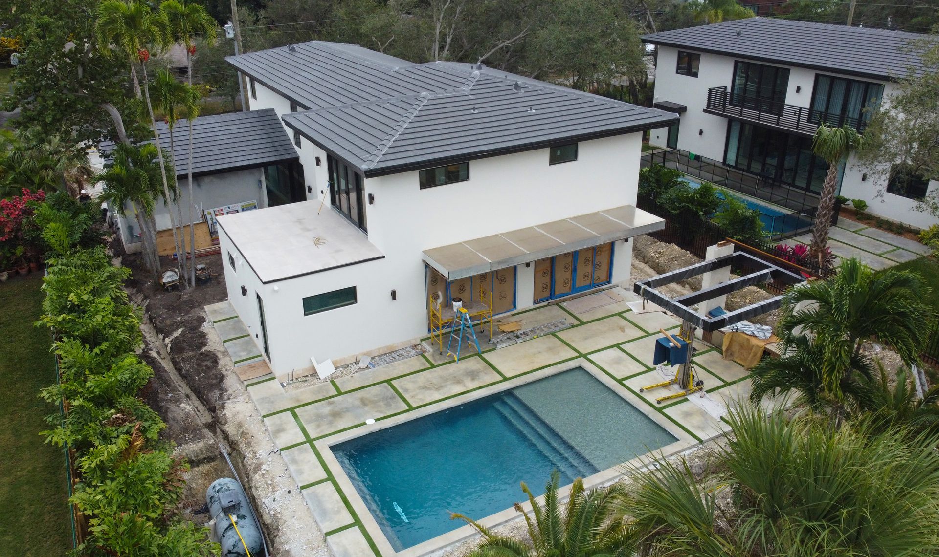 Modern white house with a pool and dark gray roof. The yard has a stone patio, and palm trees are visible.