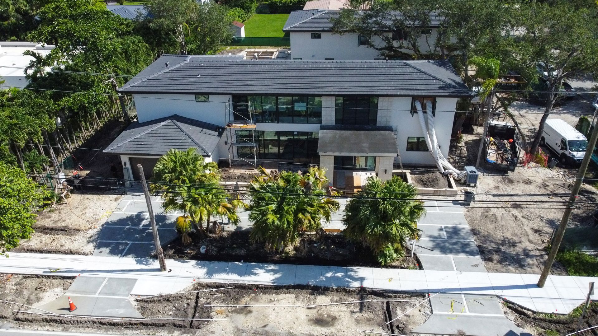 Modern two-story house with dark roof and white exterior, under construction. Trees, driveway, and street in foreground.