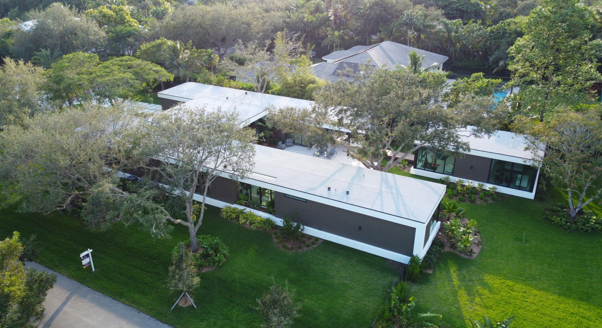 Aerial view of modern, multi-sectioned home with white roof, surrounded by green grass and trees.