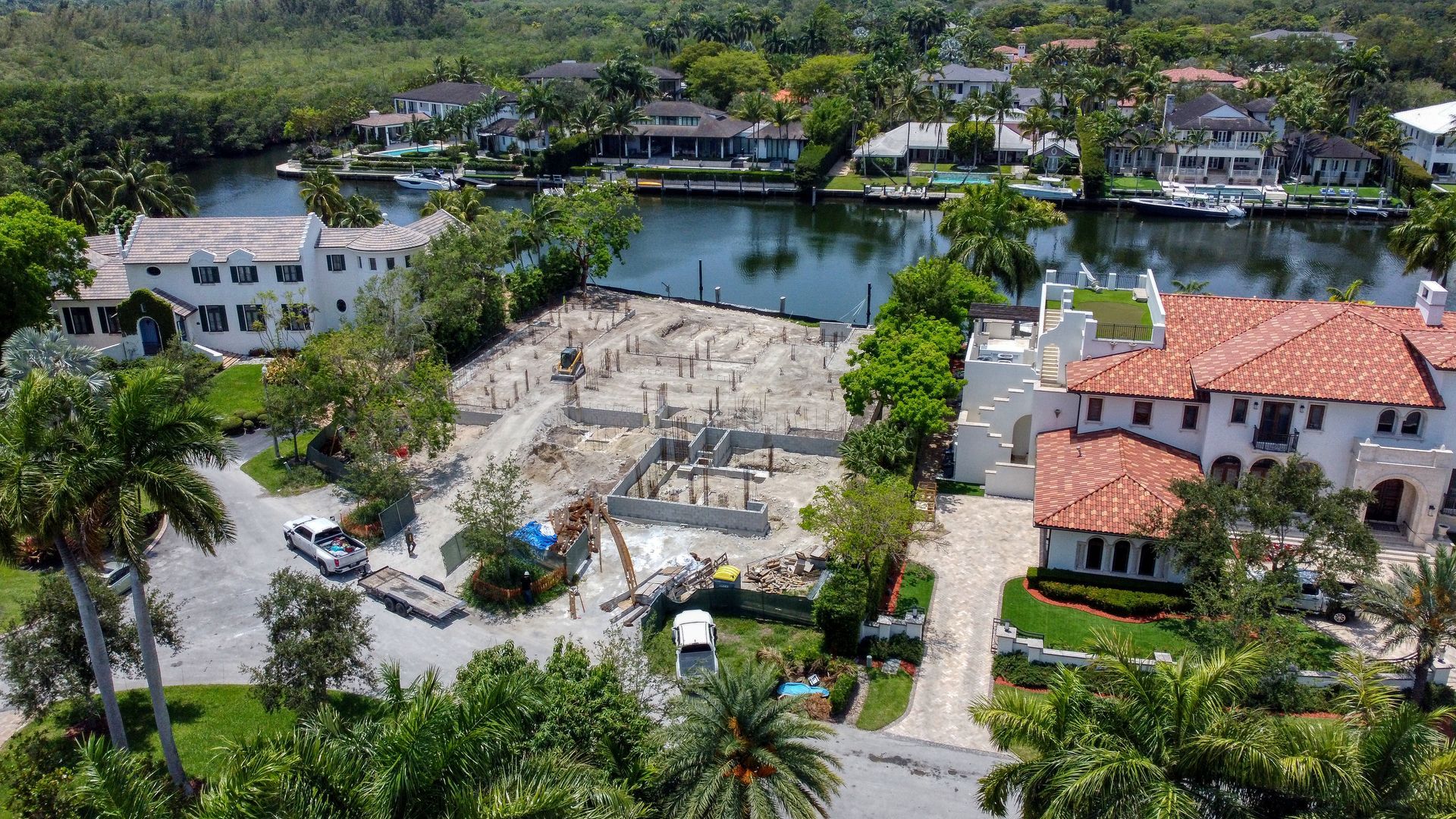 Construction site between waterfront homes; workers, equipment, foundation, waterway visible.