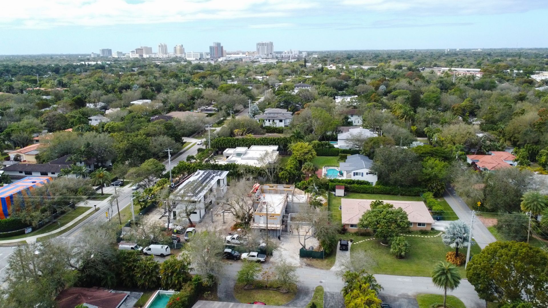Aerial view of a suburban neighborhood with houses, lush trees, a gas station, and a city skyline in the distance.