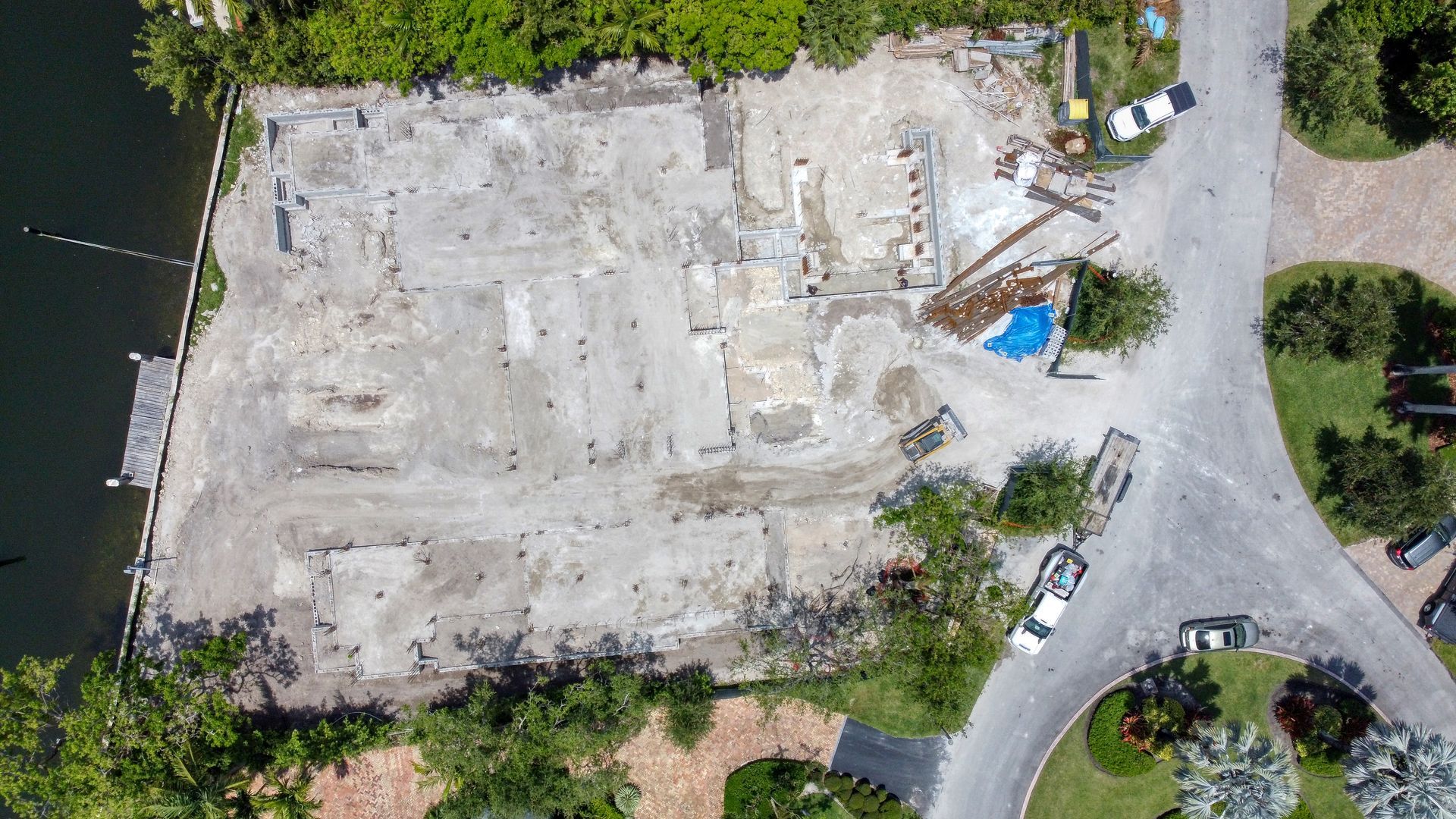 Overhead view of a construction site with remnants of a foundation near water and a curved road with cars.