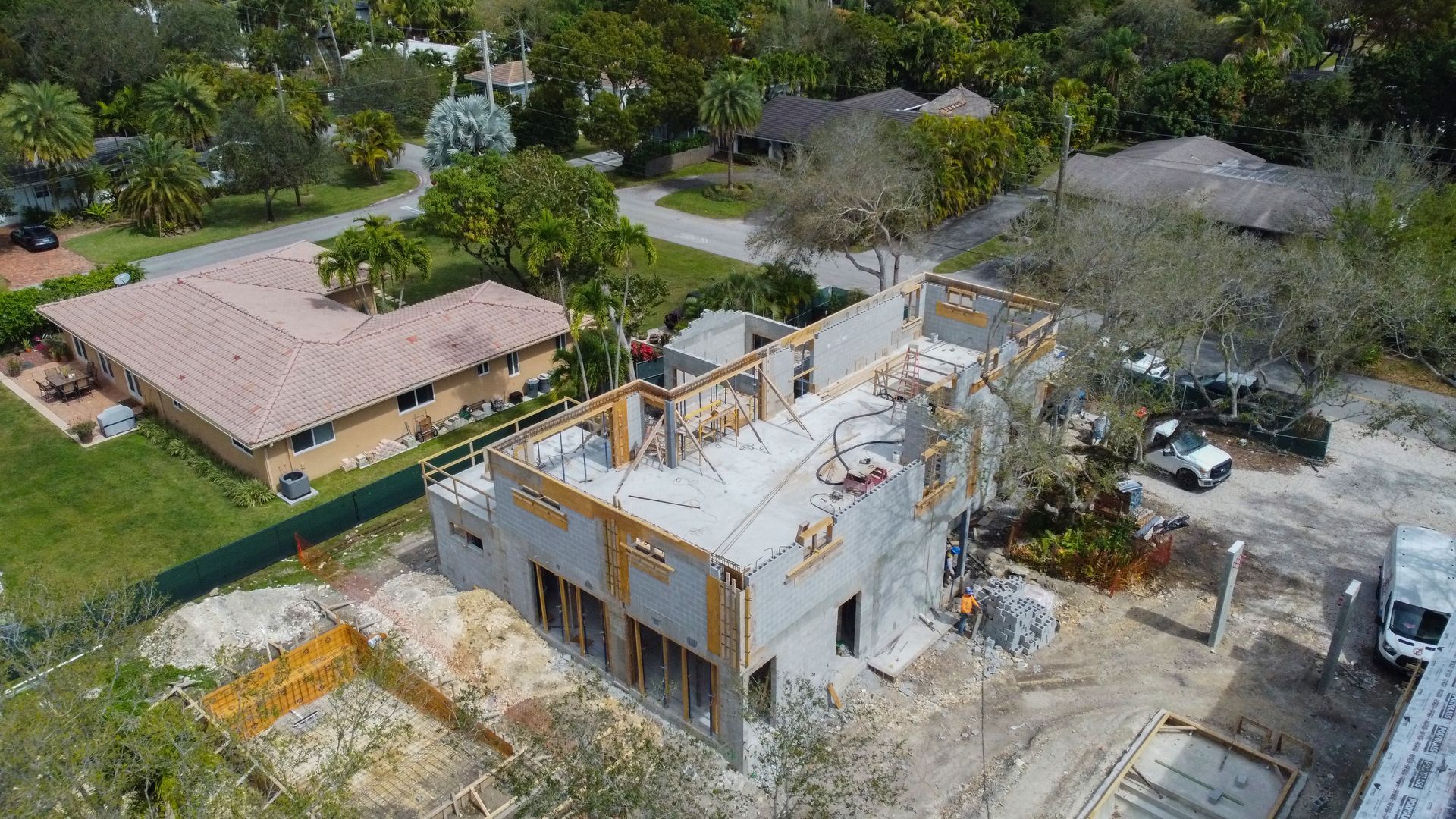 Aerial view of a home under construction; concrete blocks, wooden beams, surrounding greenery, and a nearby completed house.