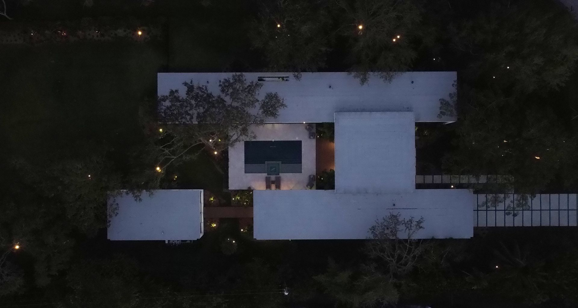 Overhead night view of a modern white house with a pool and landscaping. Small lights dot the surrounding trees.