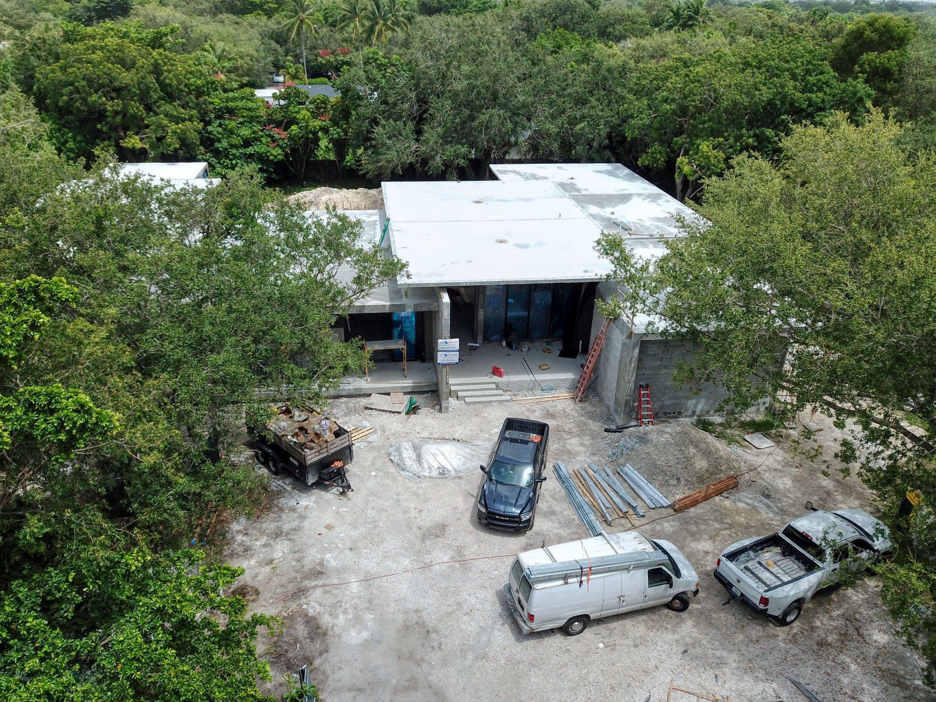 Aerial view of a building under construction, surrounded by trees. Vehicles and materials are present on the ground.