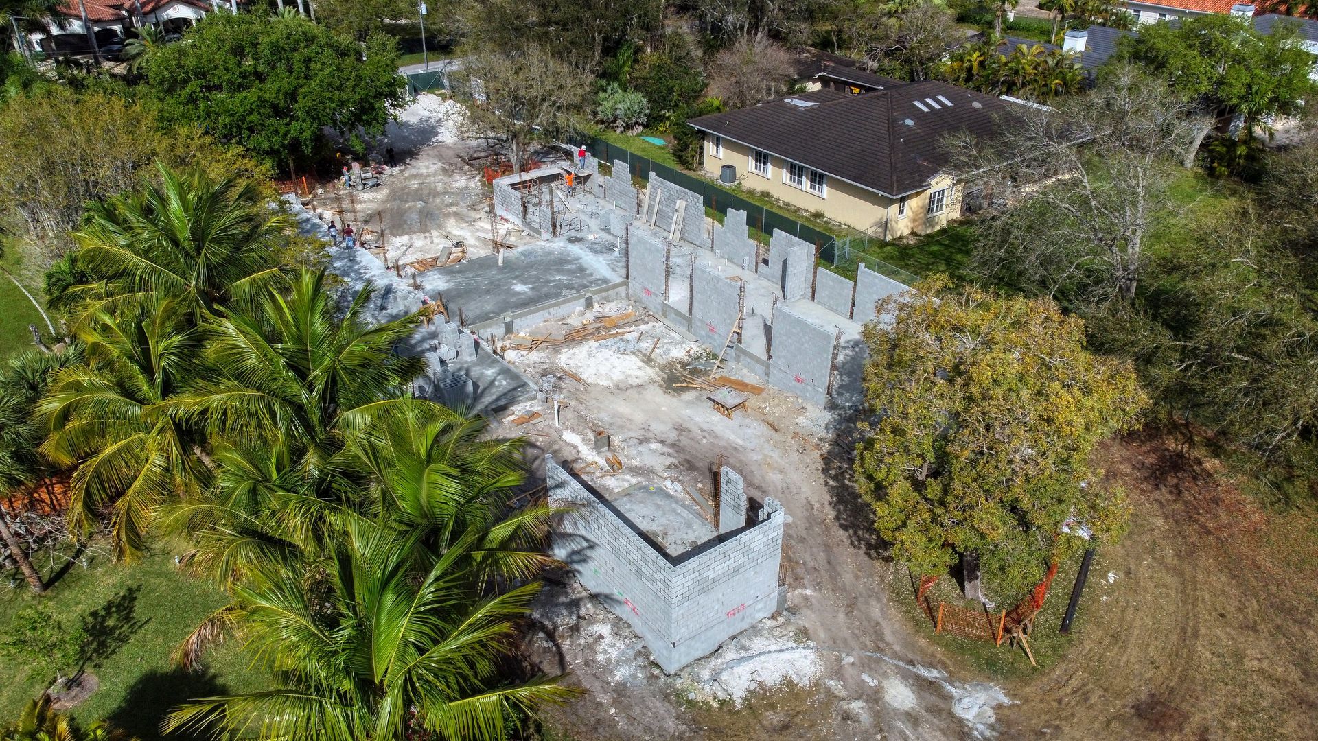 Aerial view of a construction site with concrete foundation and walls surrounded by trees and a house.