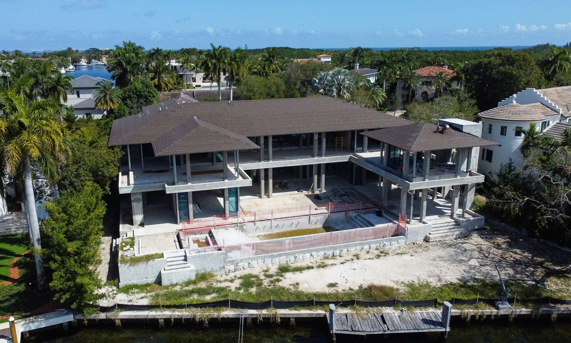 Construction of a large two-story house with a partially built pool and dock. Green trees and blue sky.