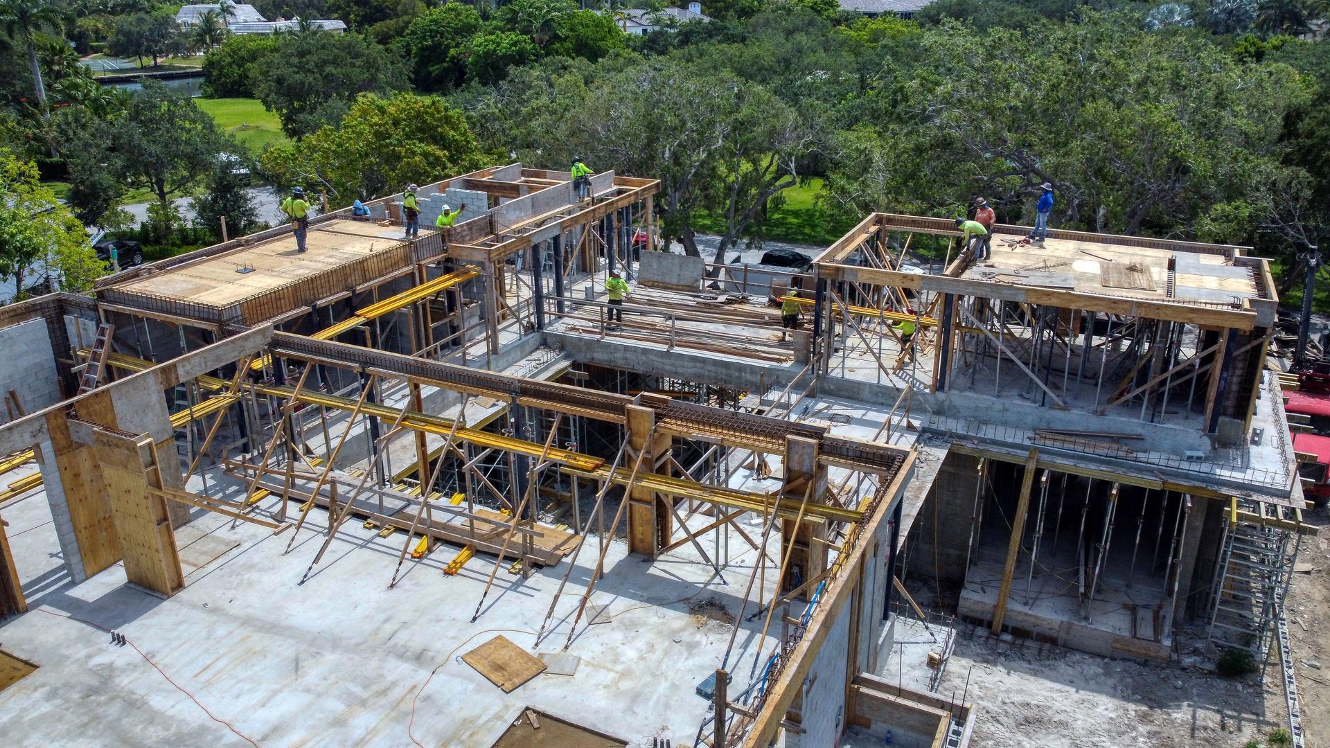 Construction site with workers, wooden framing, and concrete structures. Trees in the background.