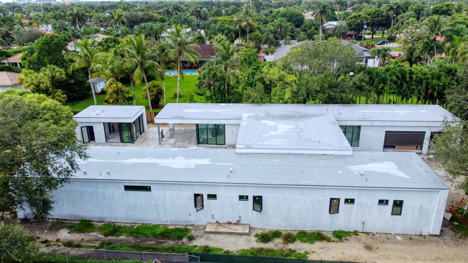 Aerial view of a long, white house with a flat roof under construction, surrounded by trees.