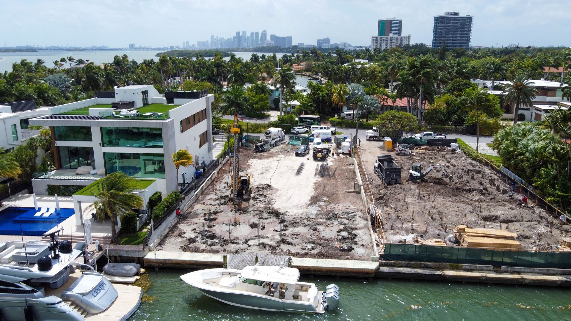 Construction site next to a waterway, with heavy machinery on dirt. Buildings and cityscape in background.