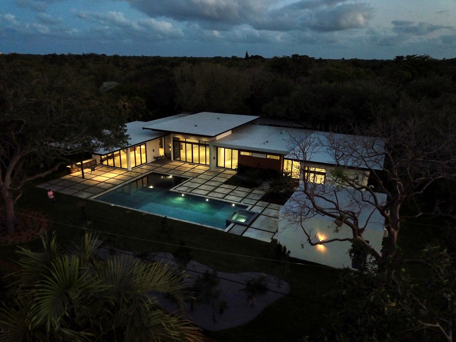 Modern house at dusk with pool, surrounded by trees. Illuminated windows, tiled patio.