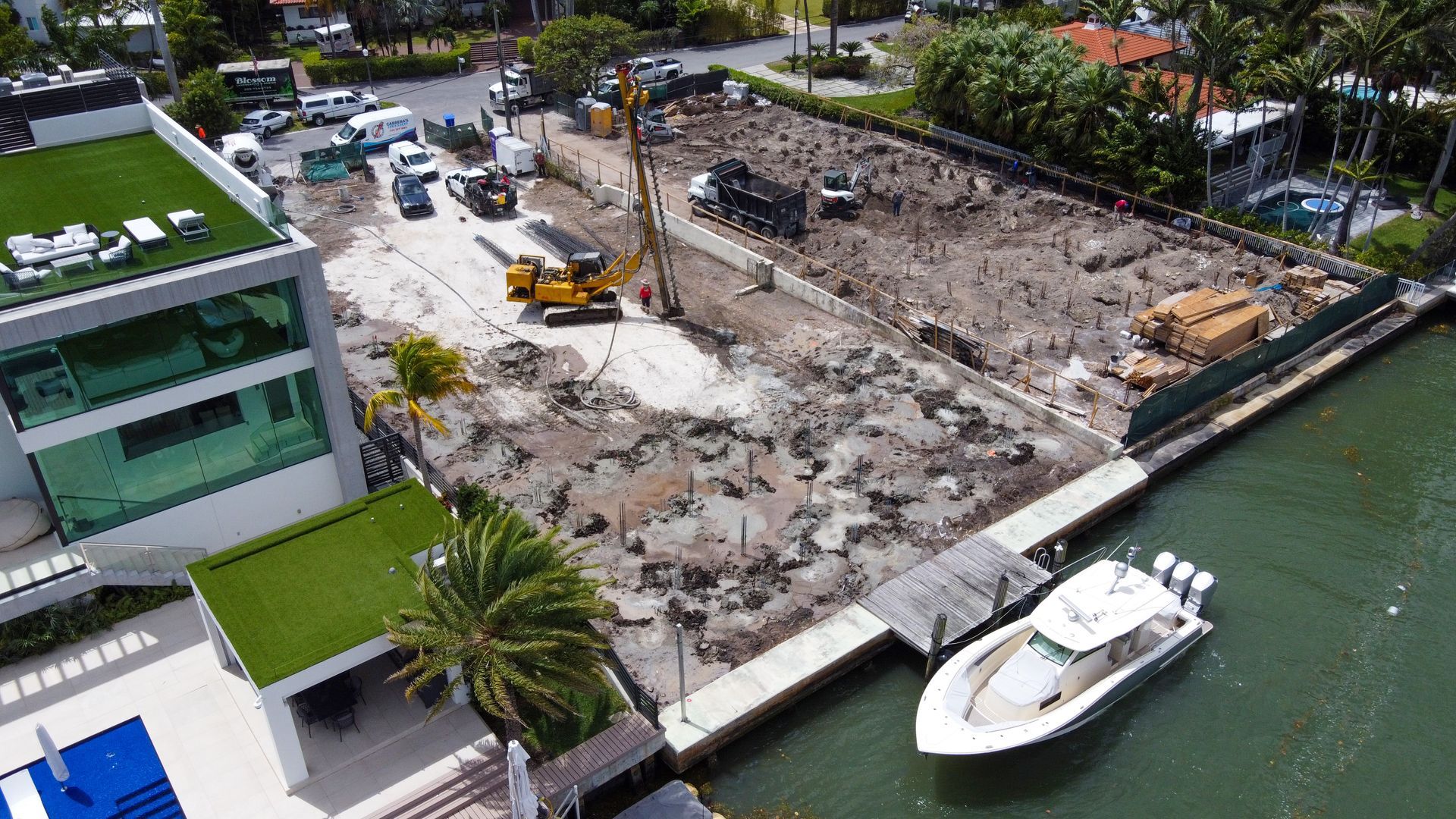 Construction site next to a building and water. Yellow construction equipment, white boat at dock.