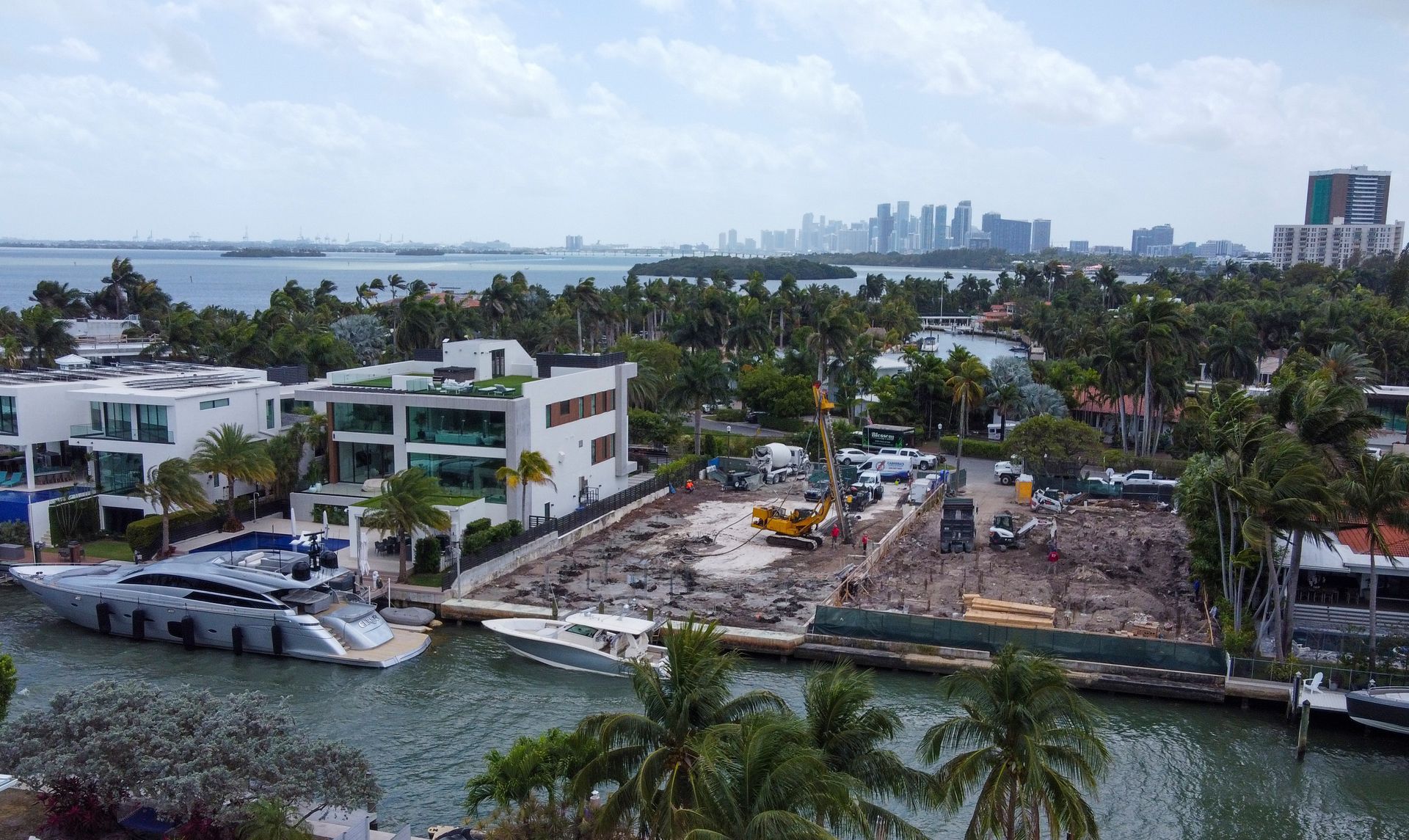 Construction site with machinery, luxury homes, yacht, water, and city skyline in the background.