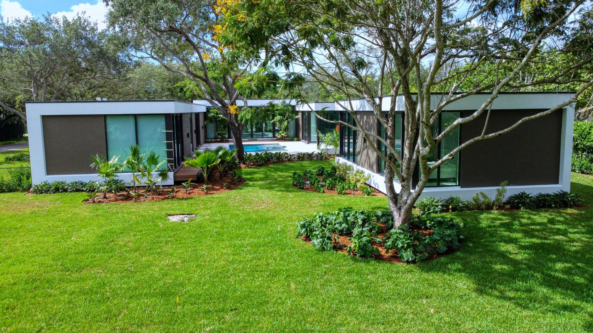 Modern, rectangular house with glass windows, brown panels, and a lush green lawn, trees, and blue water in the background.