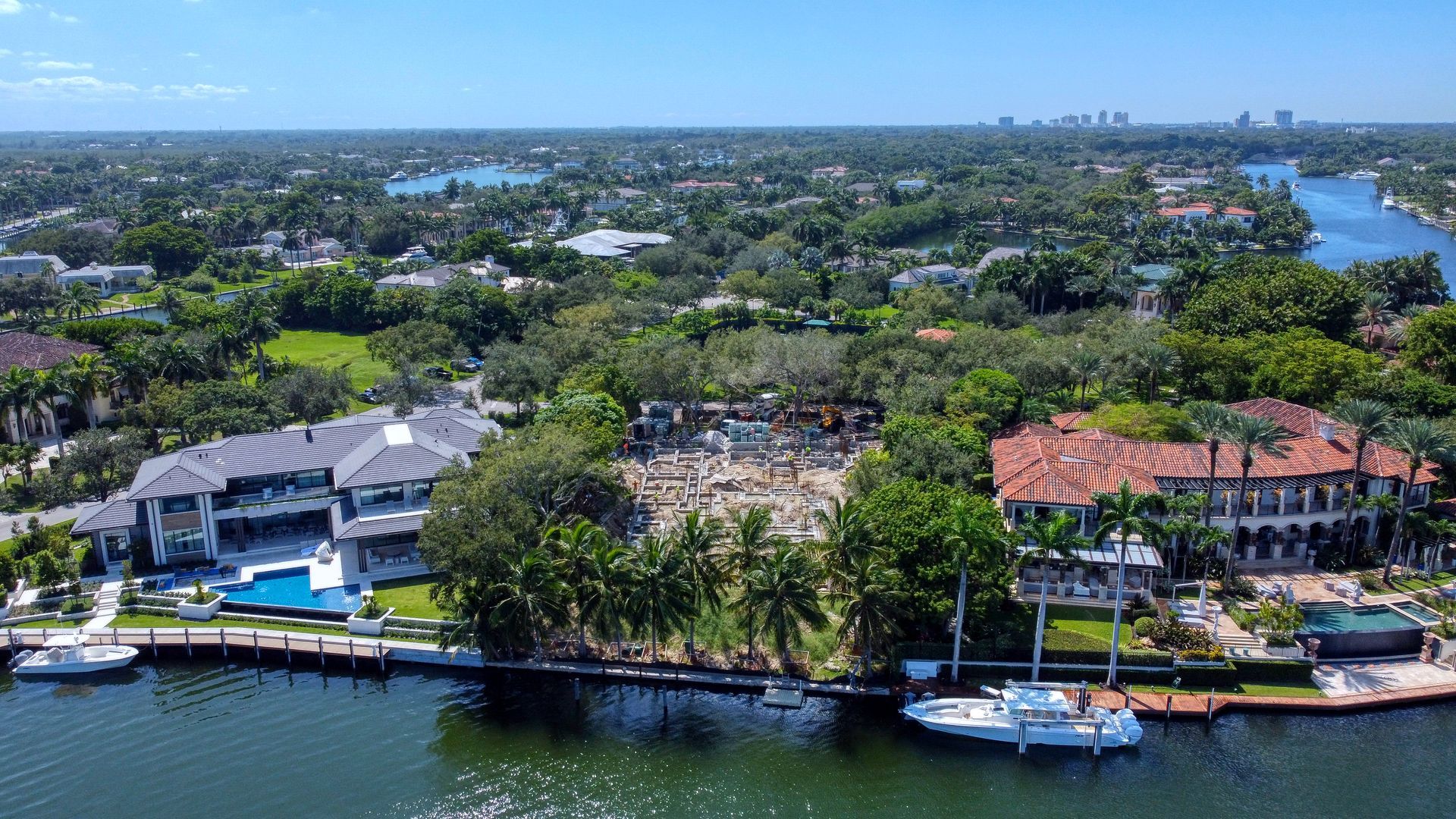 Aerial view of waterfront houses with boats and a cityscape in the background under a blue sky.