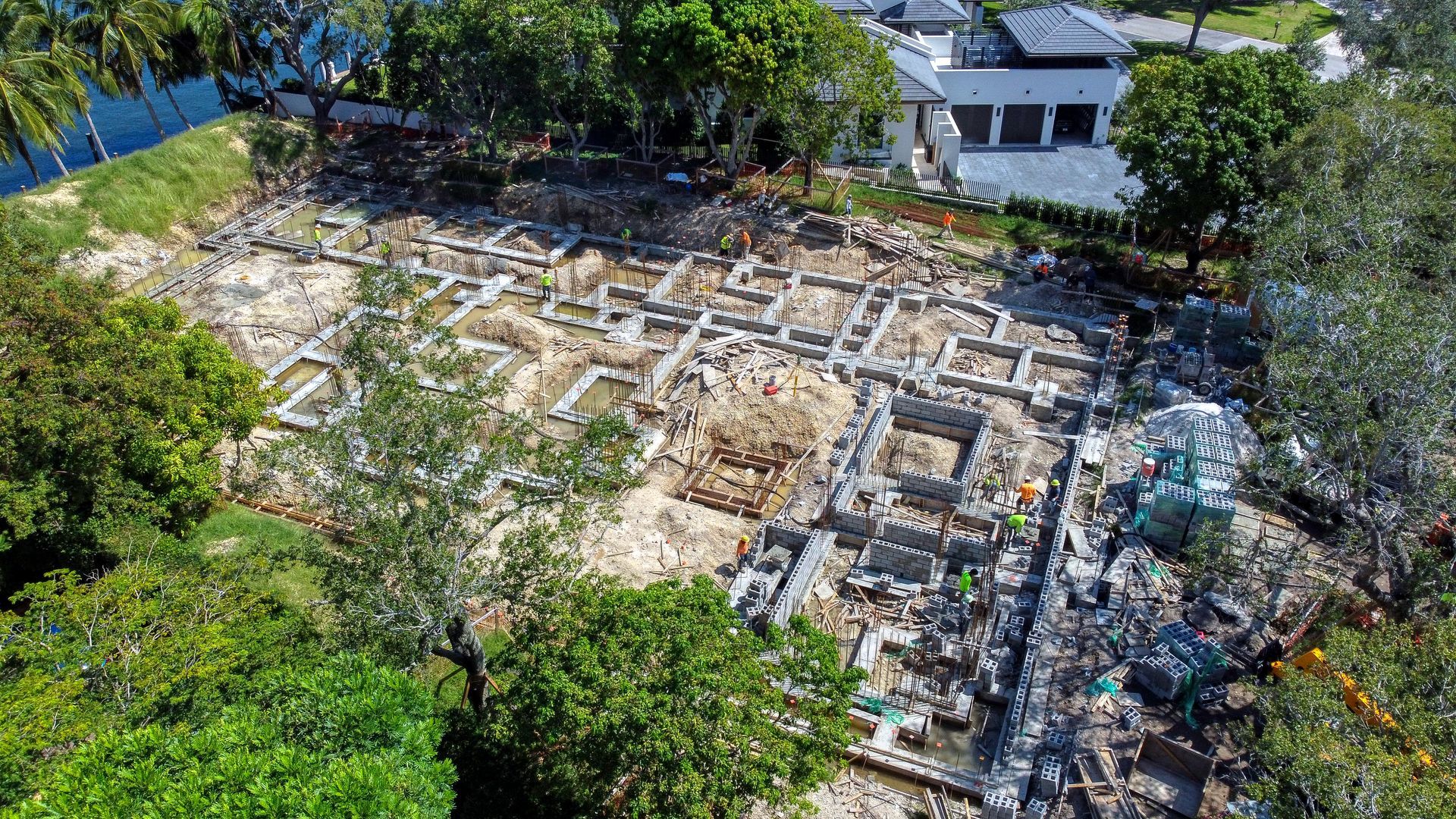 Aerial view of an archaeological dig site with visible foundation outlines and workers. Trees and a building surround.