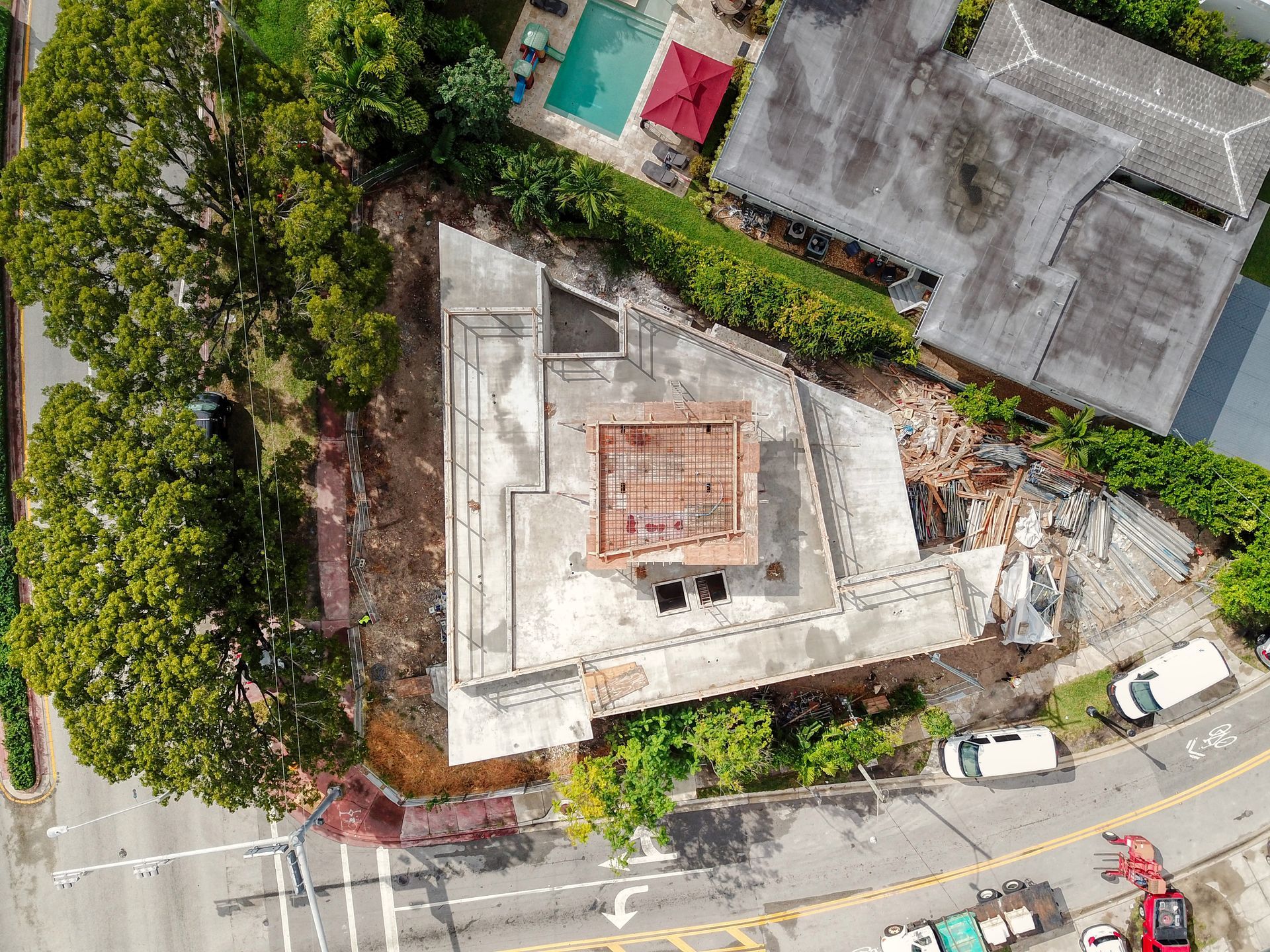 Overhead view of a partially constructed building on a triangular lot; trees, street, and a pool are also visible.