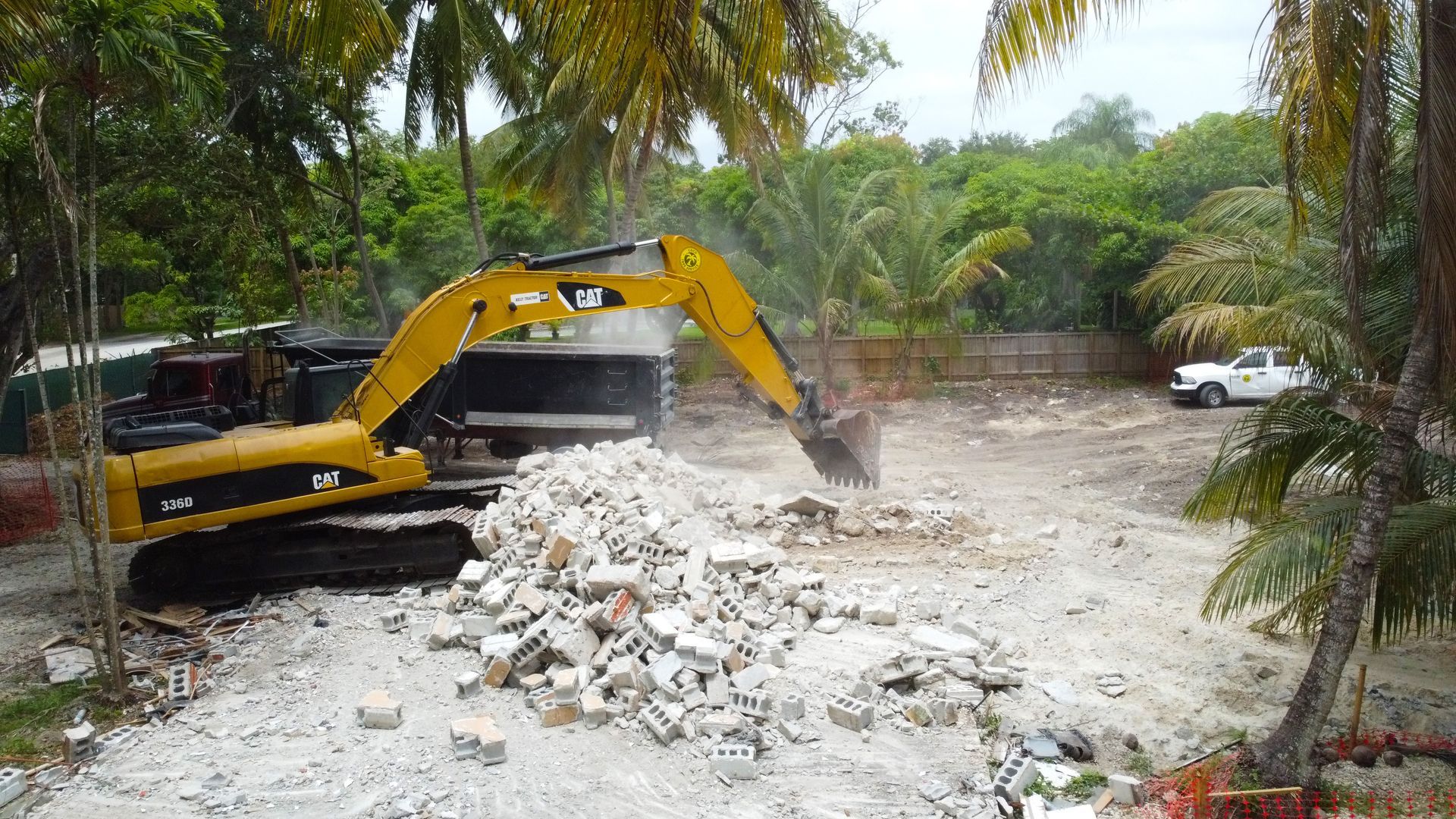 Yellow excavator demolishes a building, surrounded by rubble, trees, and a white truck.
