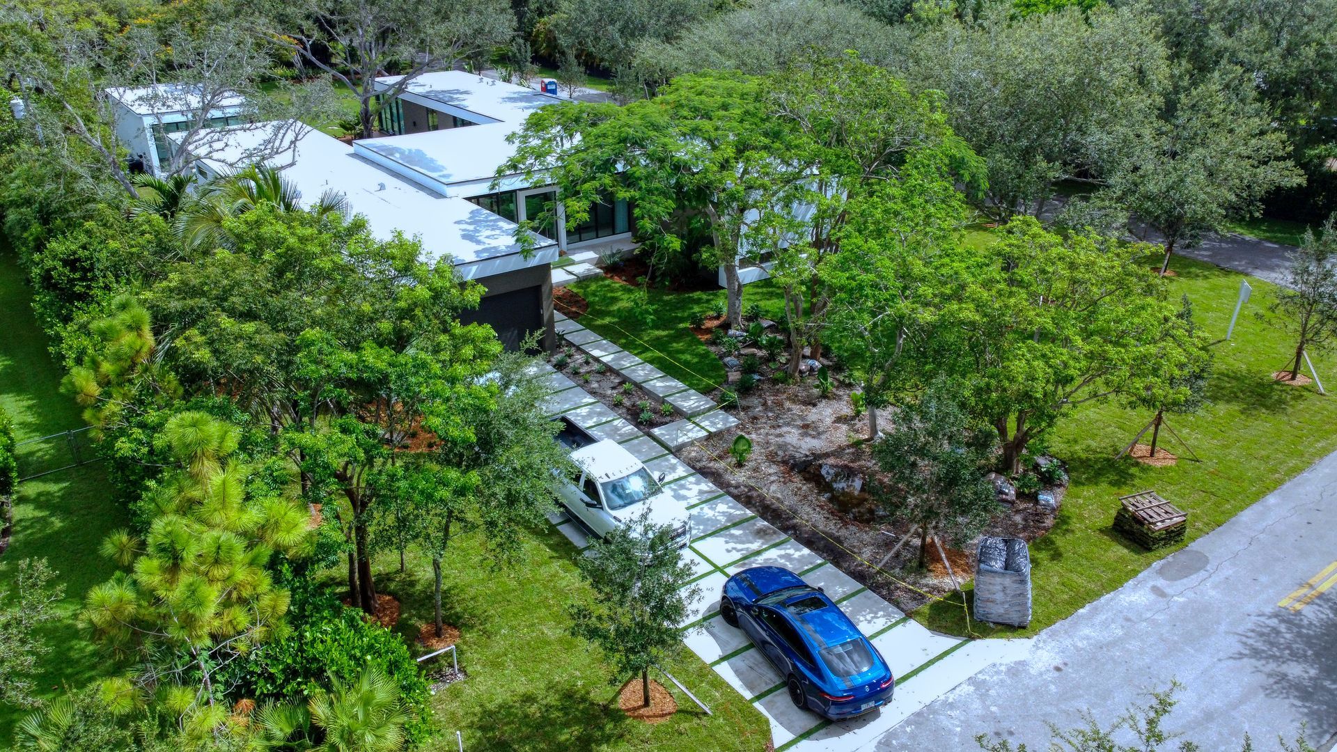 Aerial view of a modern home with a long driveway and two cars parked. Green trees surround the property.