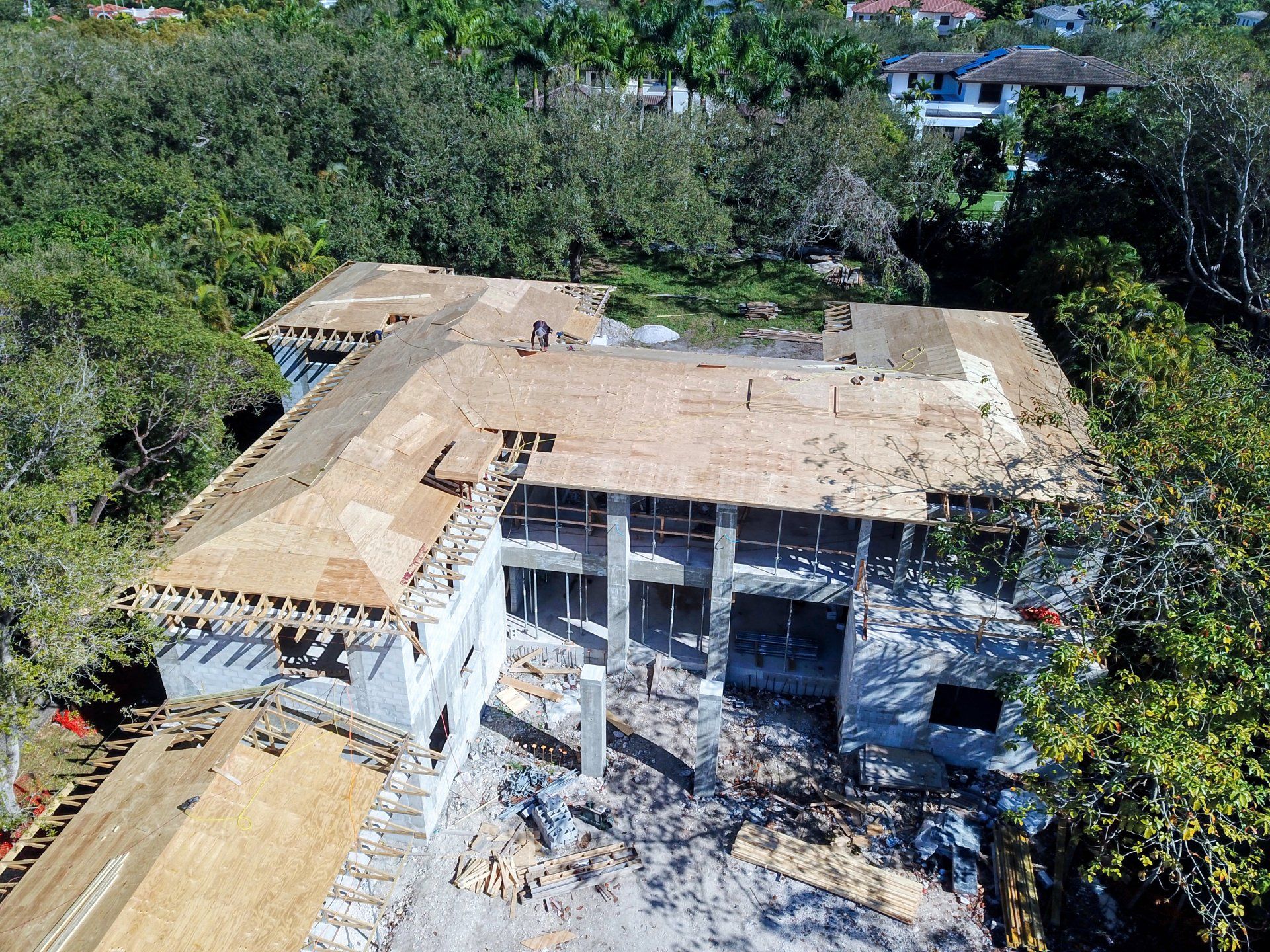 Aerial view of a house under construction with exposed roof framing and partially constructed walls surrounded by trees.