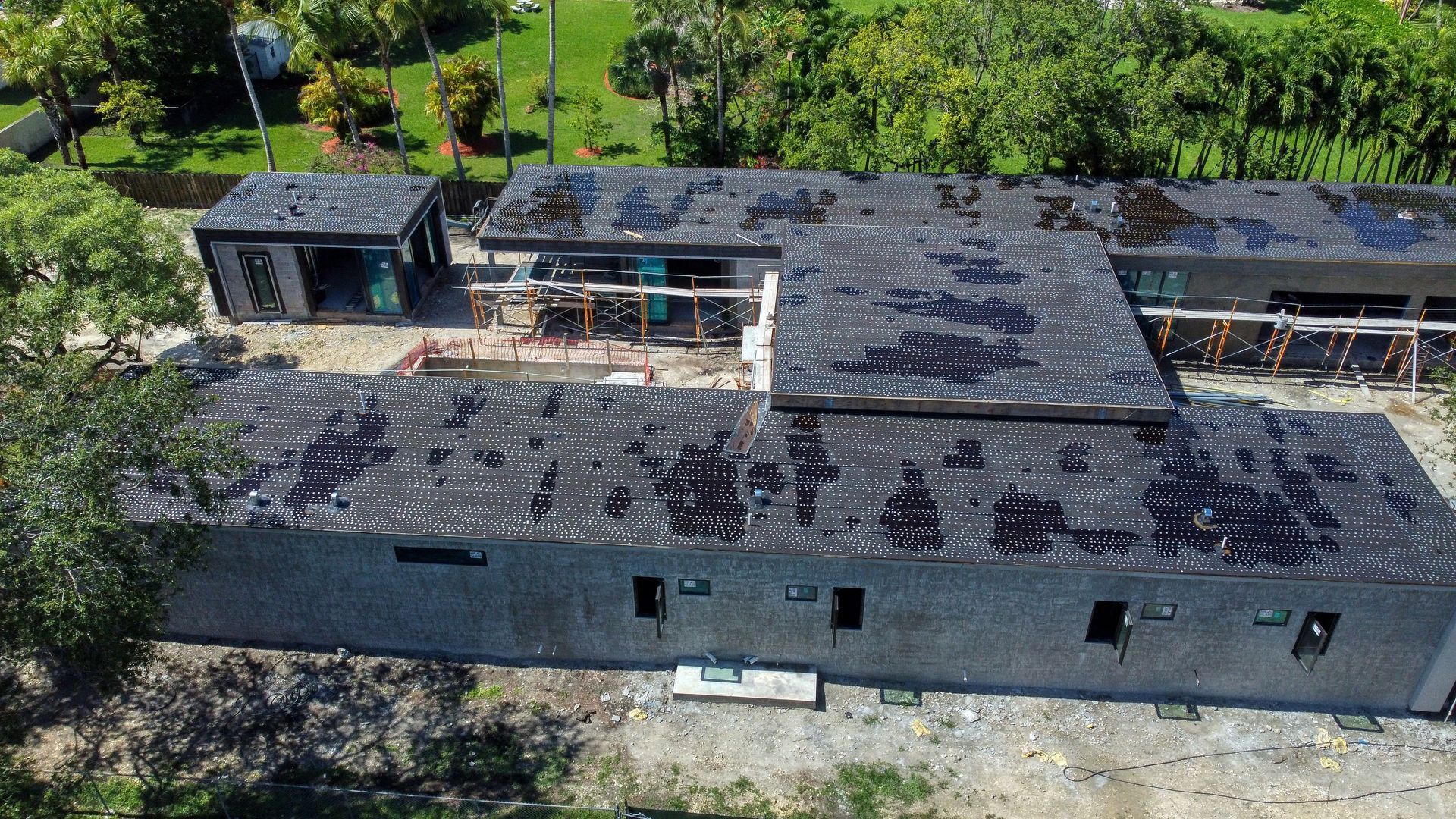 Aerial view of a long, gray building with black, textured roofing. Trees and grass surround it.