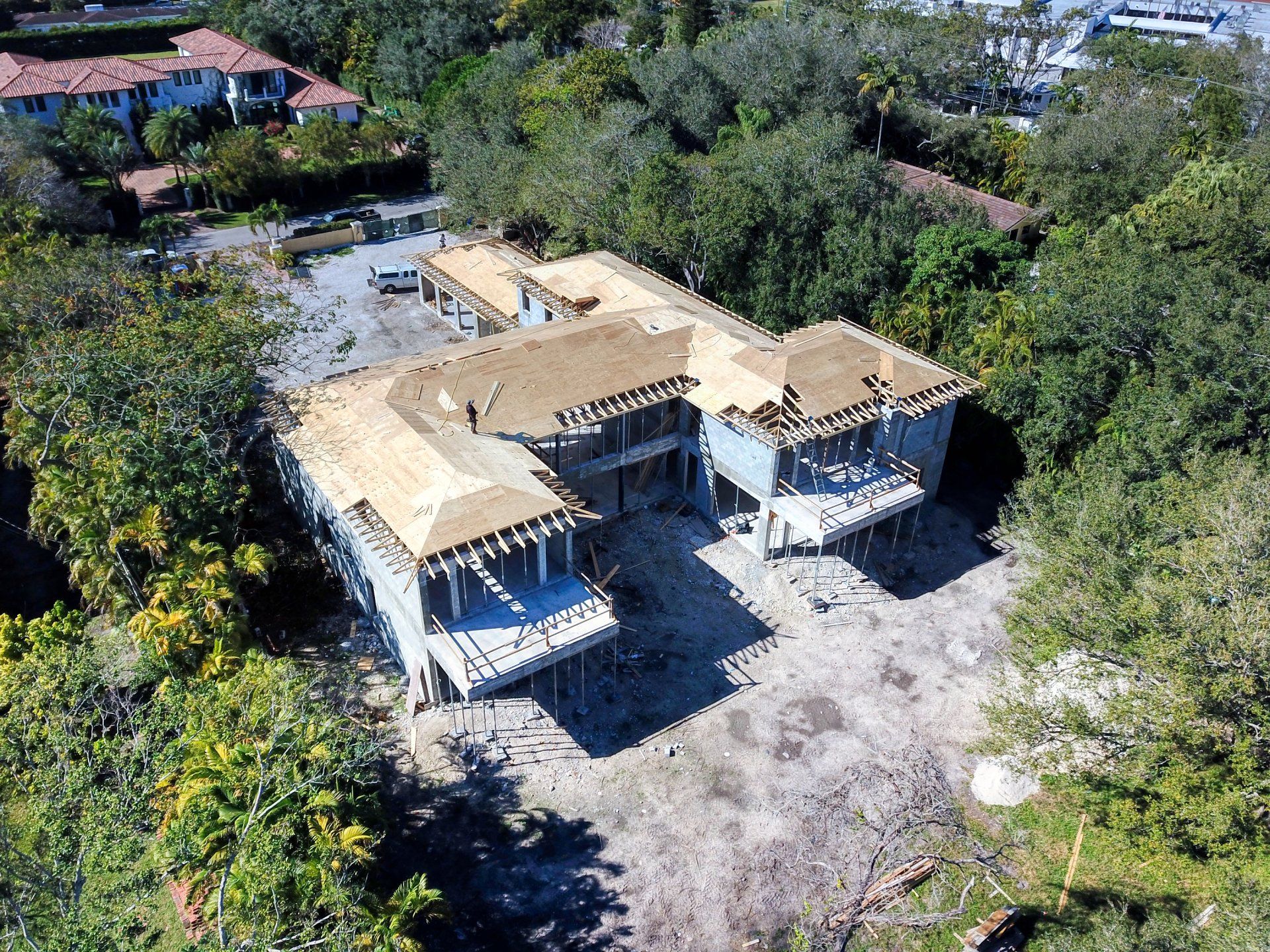 Aerial view of a house under construction with exposed framing and a partially completed roof, surrounded by trees.