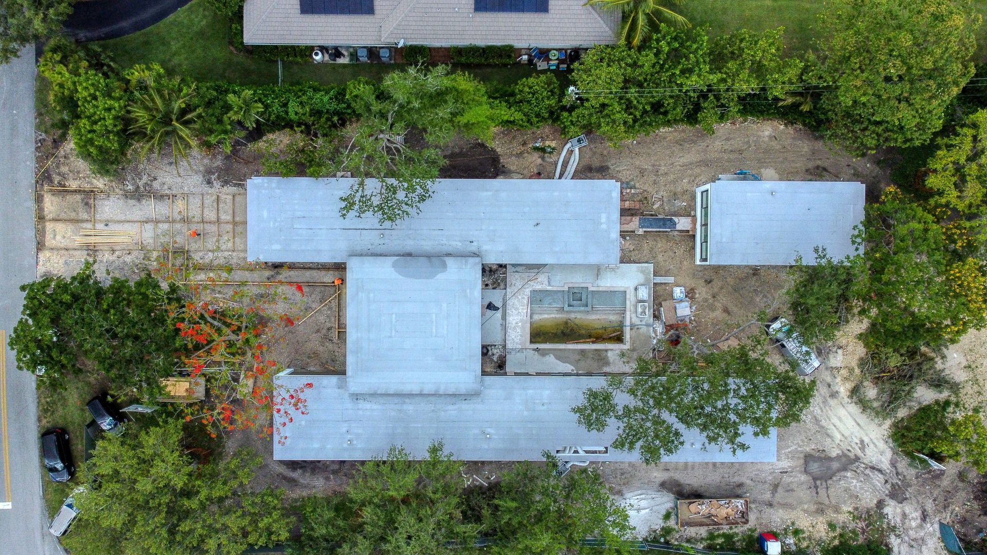 Aerial view of a building under construction, featuring gray rooftops, surrounded by trees and construction materials.