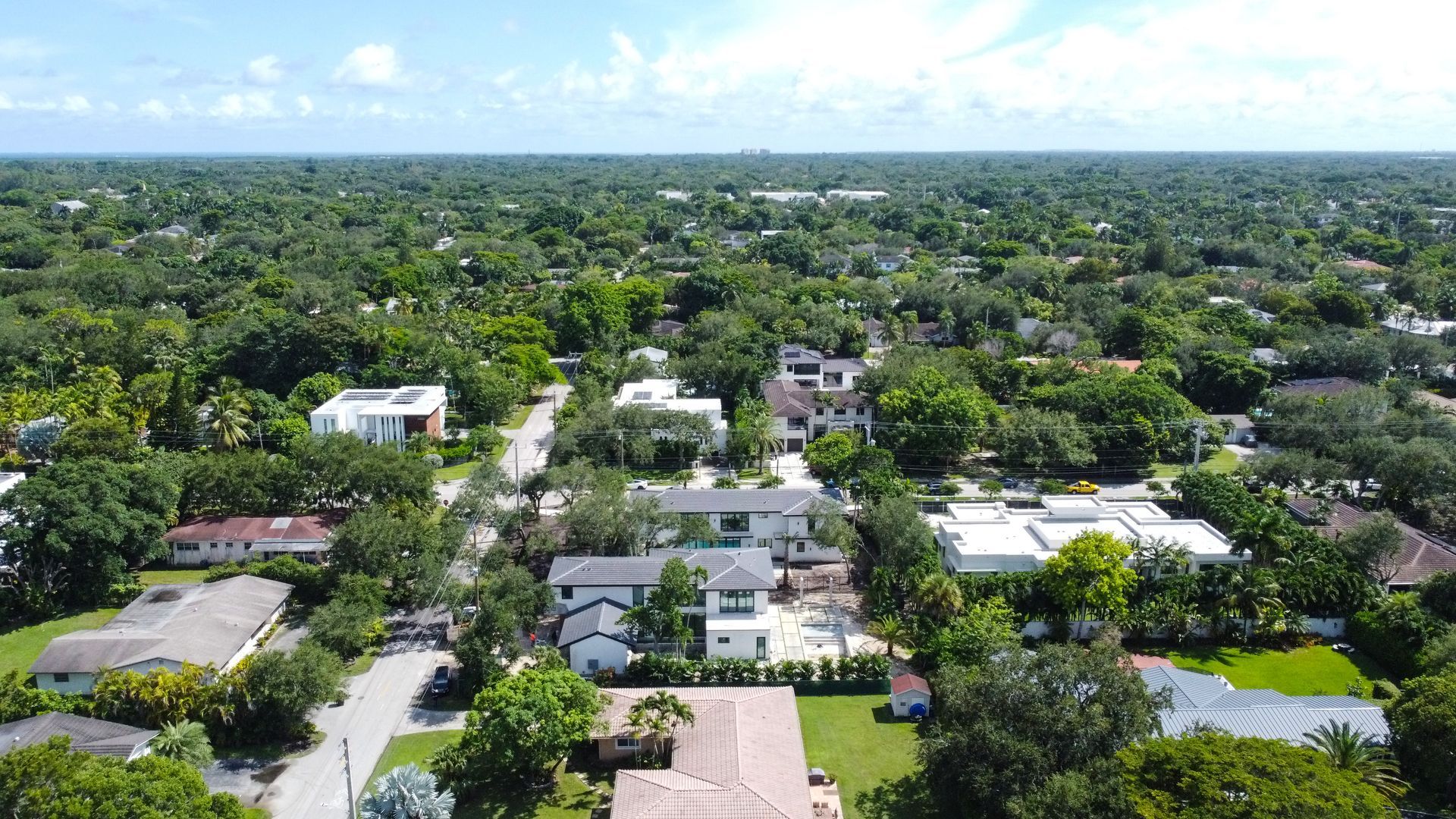 Aerial view of a residential area with houses surrounded by lush green trees under a bright sky.