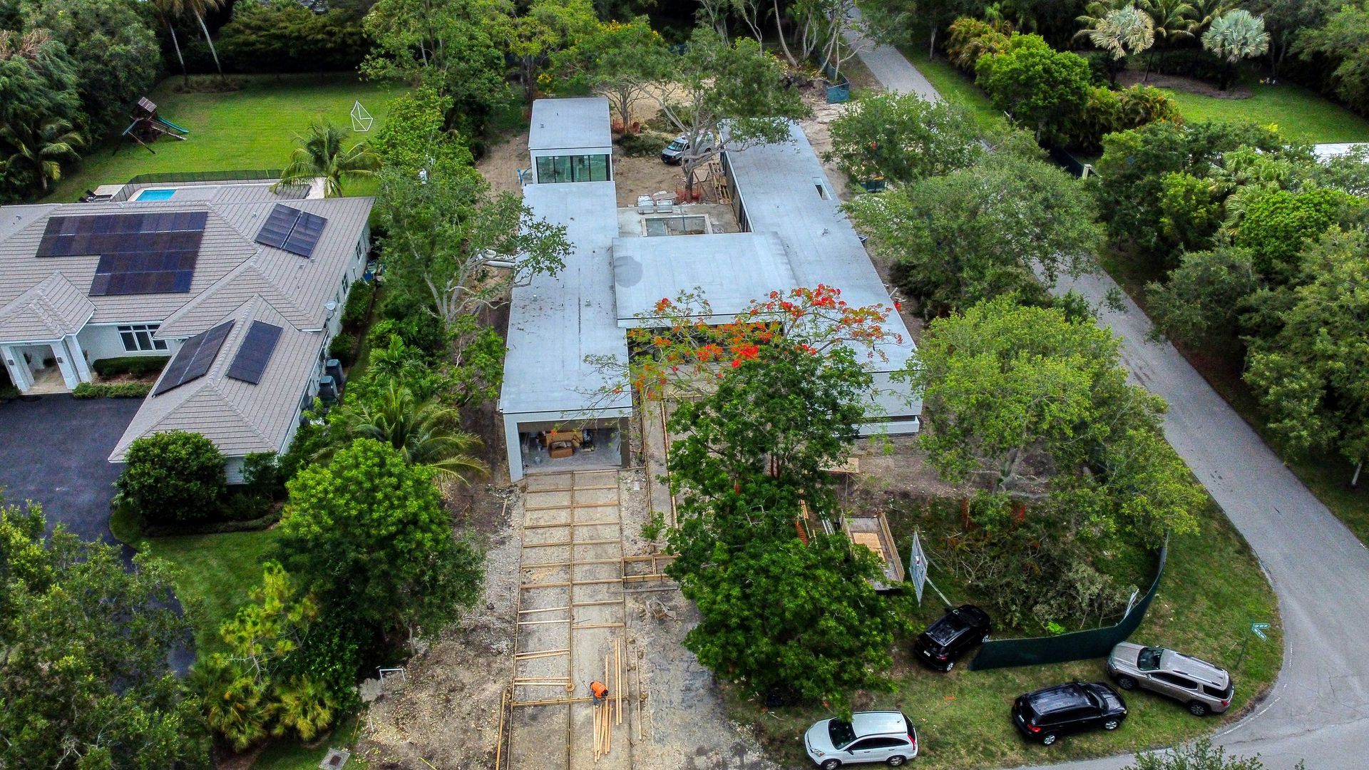 Aerial view of a long, light-colored building with a gray roof and numerous surrounding trees and vehicles.