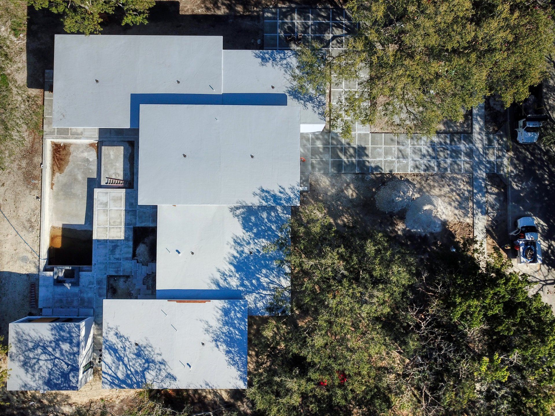 Overhead view of a modern building with multiple flat gray roofs, trees surrounding it, and parked cars.