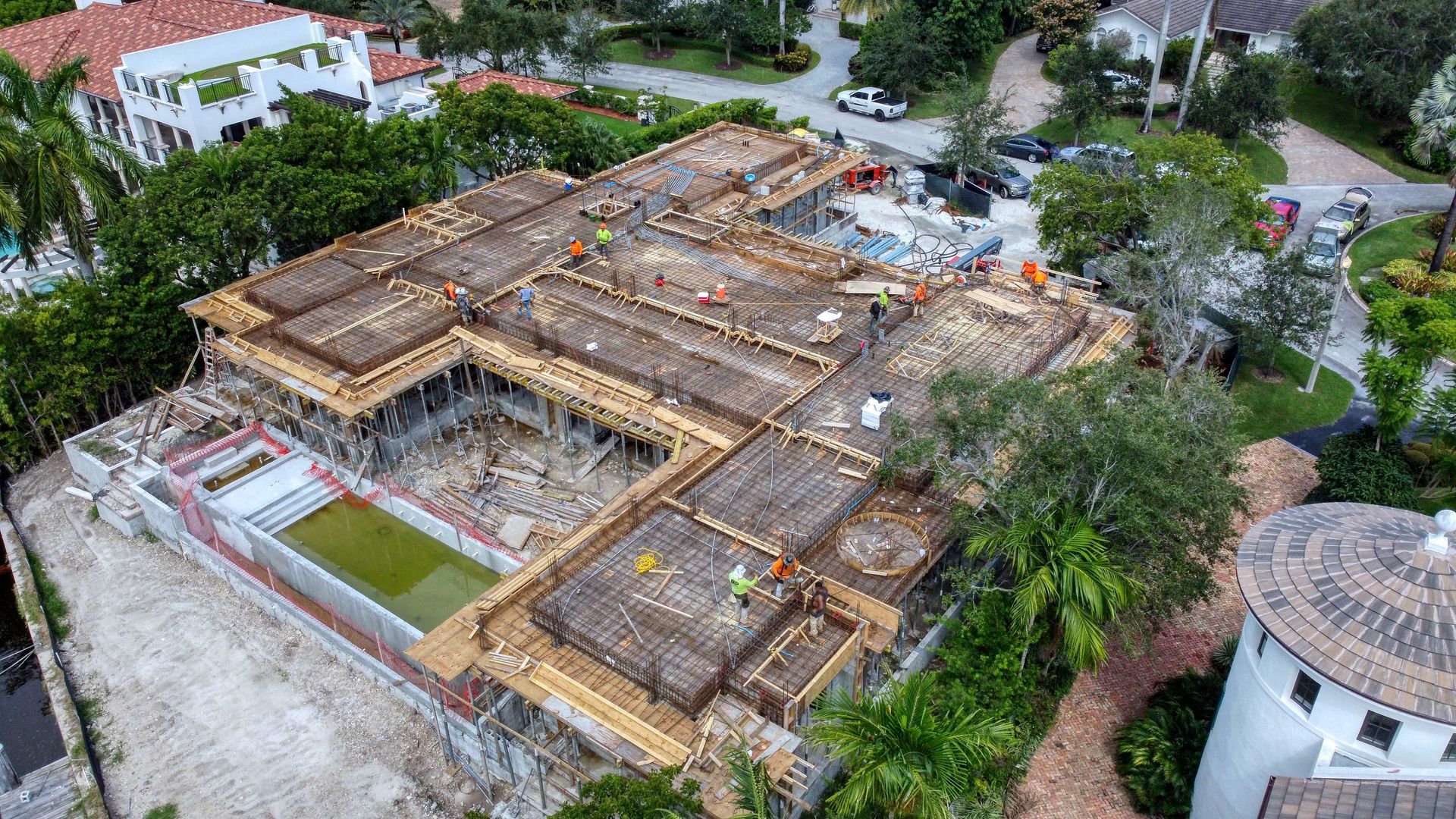 Aerial view of a large building under construction with exposed beams, surrounded by trees and residential homes.
