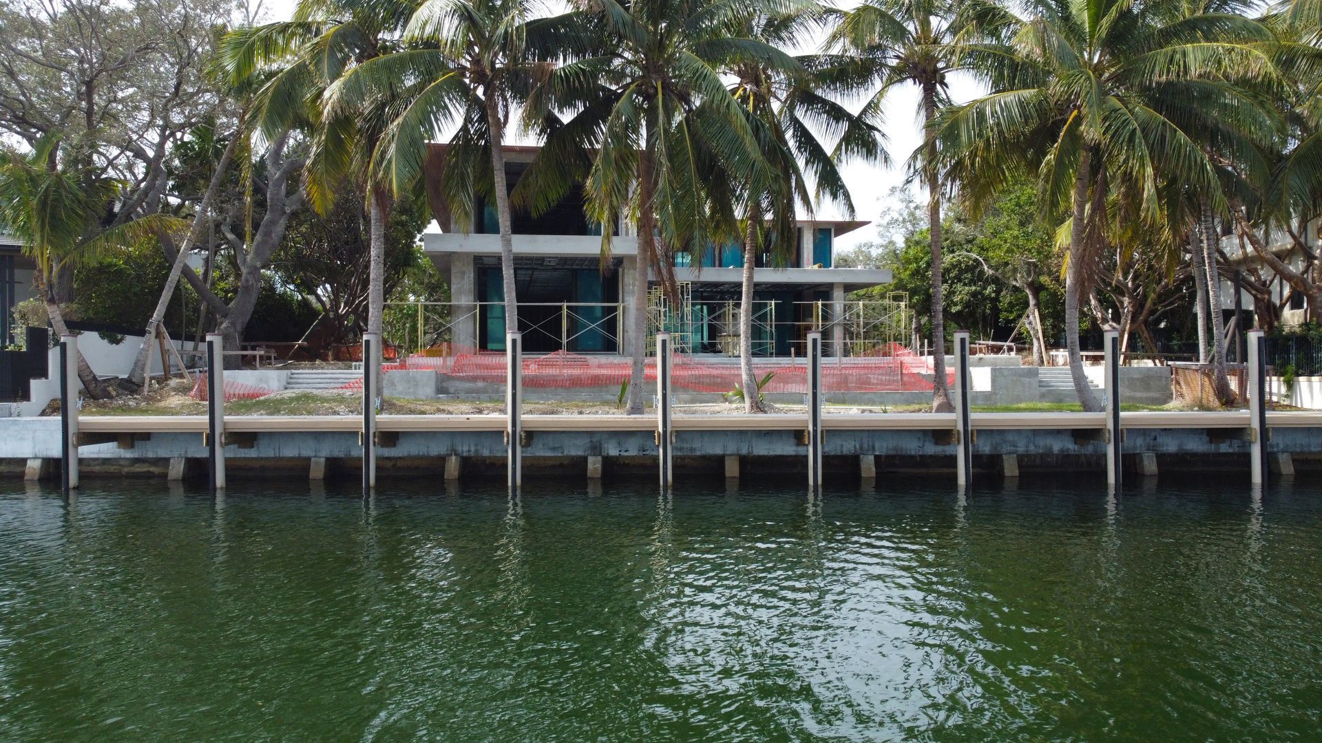 House under construction behind a waterfront dock, with palm trees and a view of the water.