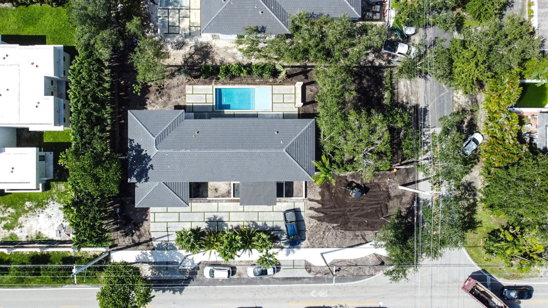 Aerial view of a house with a pool, trees, and a street. Gray roof, blue pool, brown car.