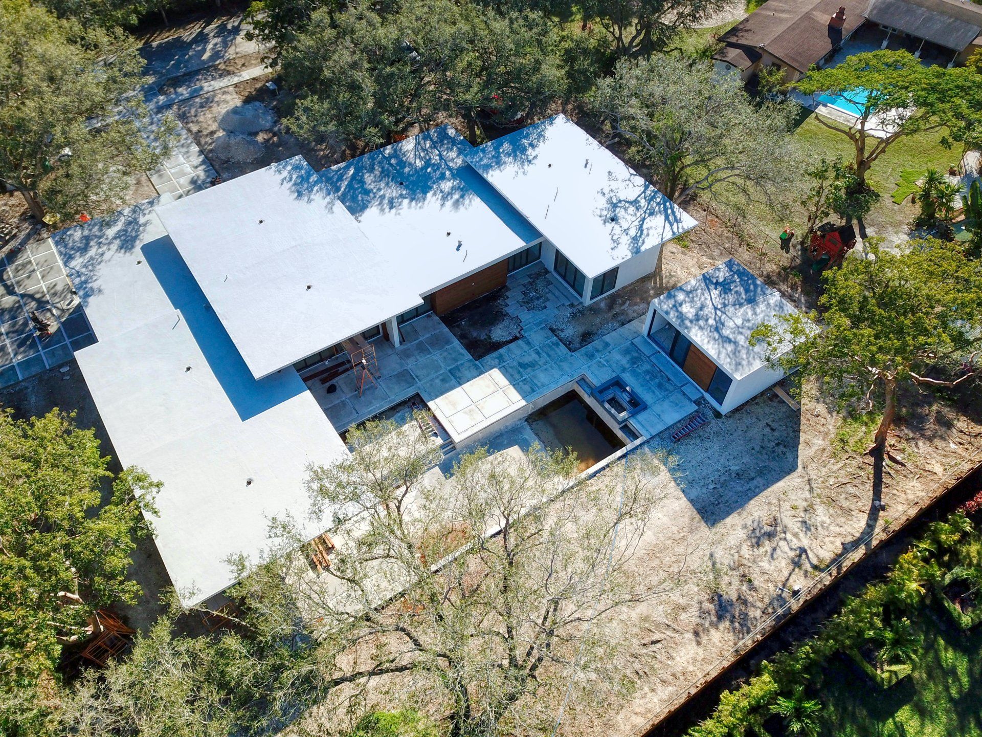 Aerial view of a modern white-roofed house surrounded by trees, with a pool and patio area.