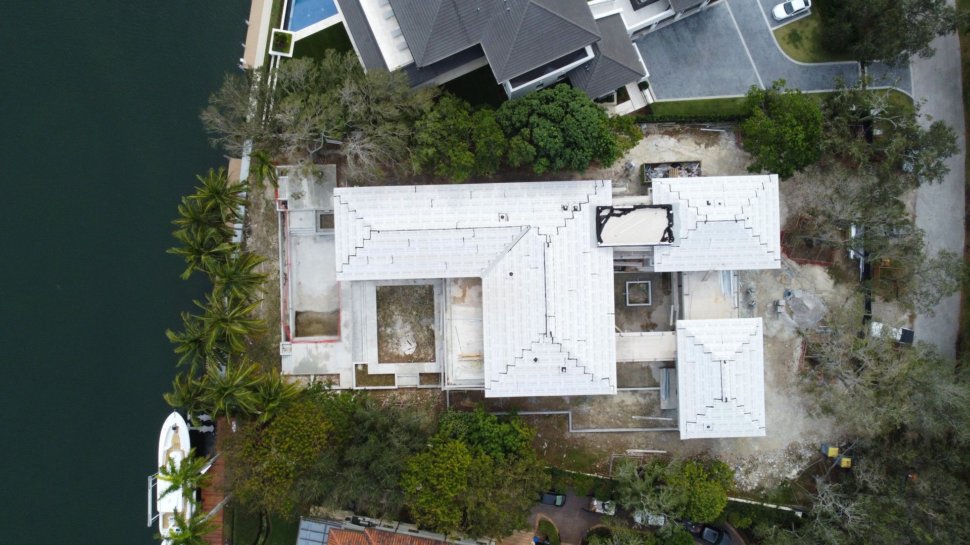 Aerial view of a house under construction with a white roof near a body of water and trees.