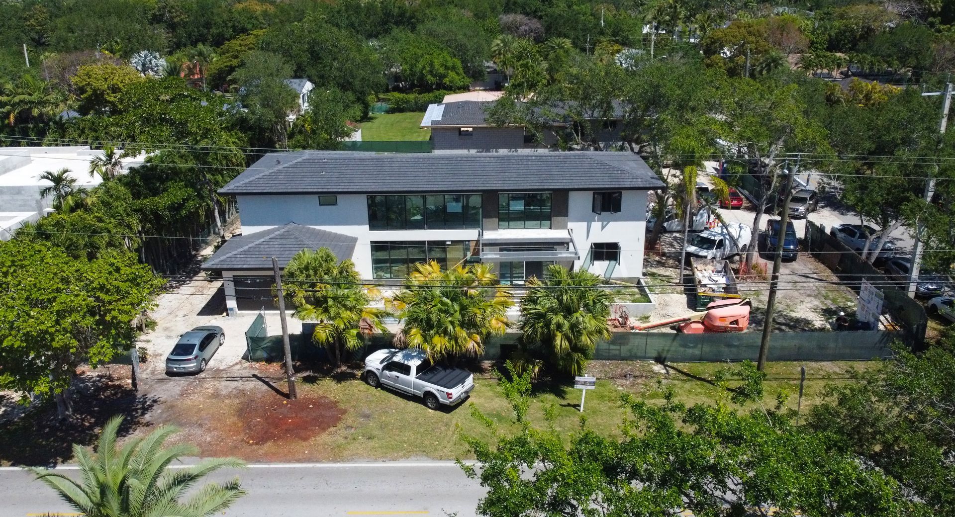 Two-story modern home with dark roof, surrounded by trees, construction equipment, and parked vehicles.