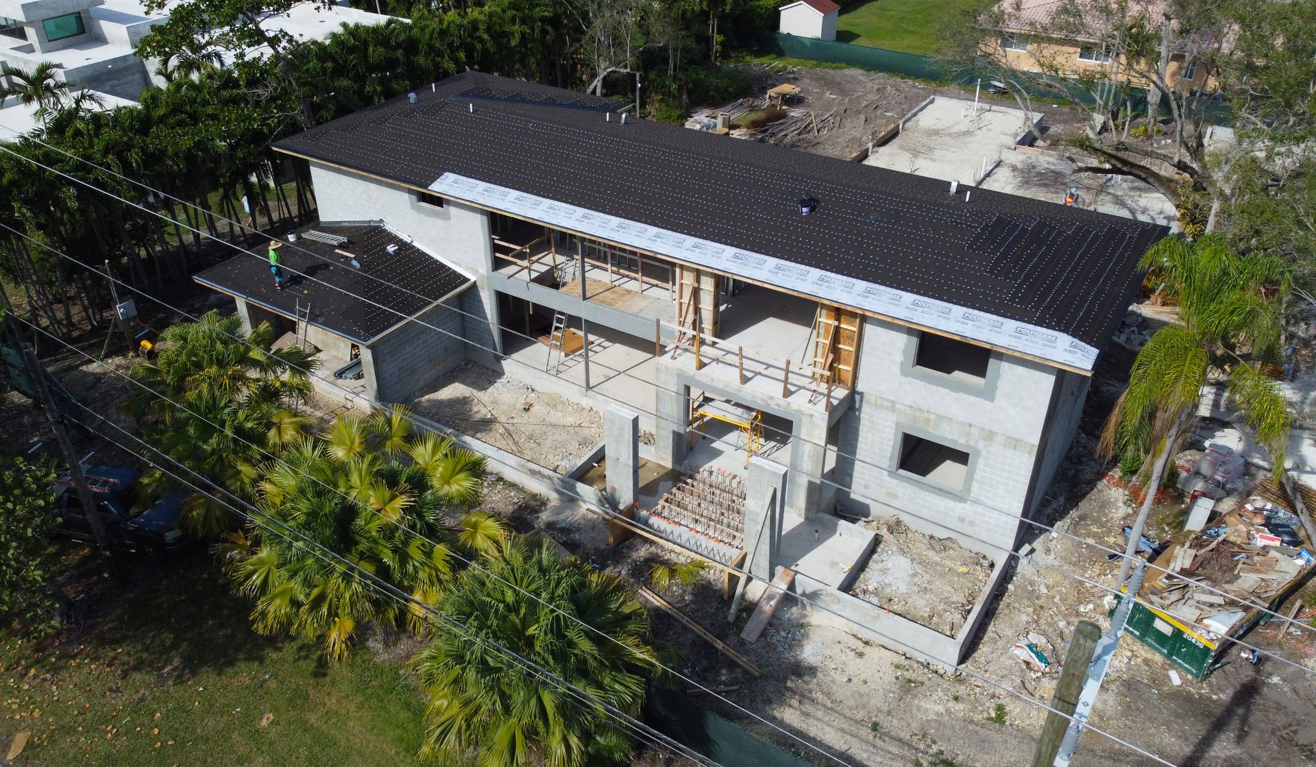 Aerial view of a two-story concrete building under construction with a dark roof, surrounded by trees and green grass.