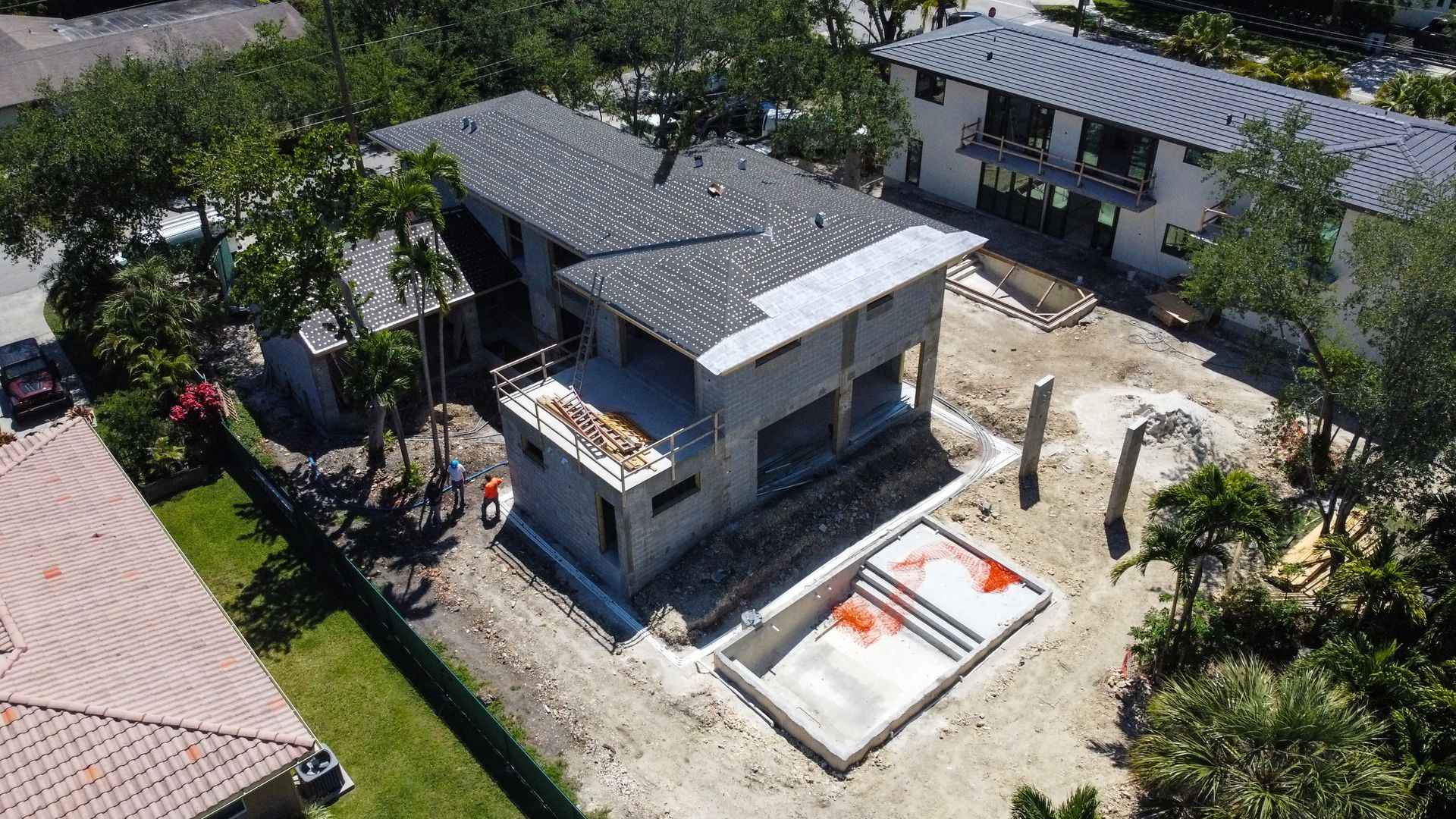 Aerial view of a house under construction with a pool, surrounded by trees and other buildings.