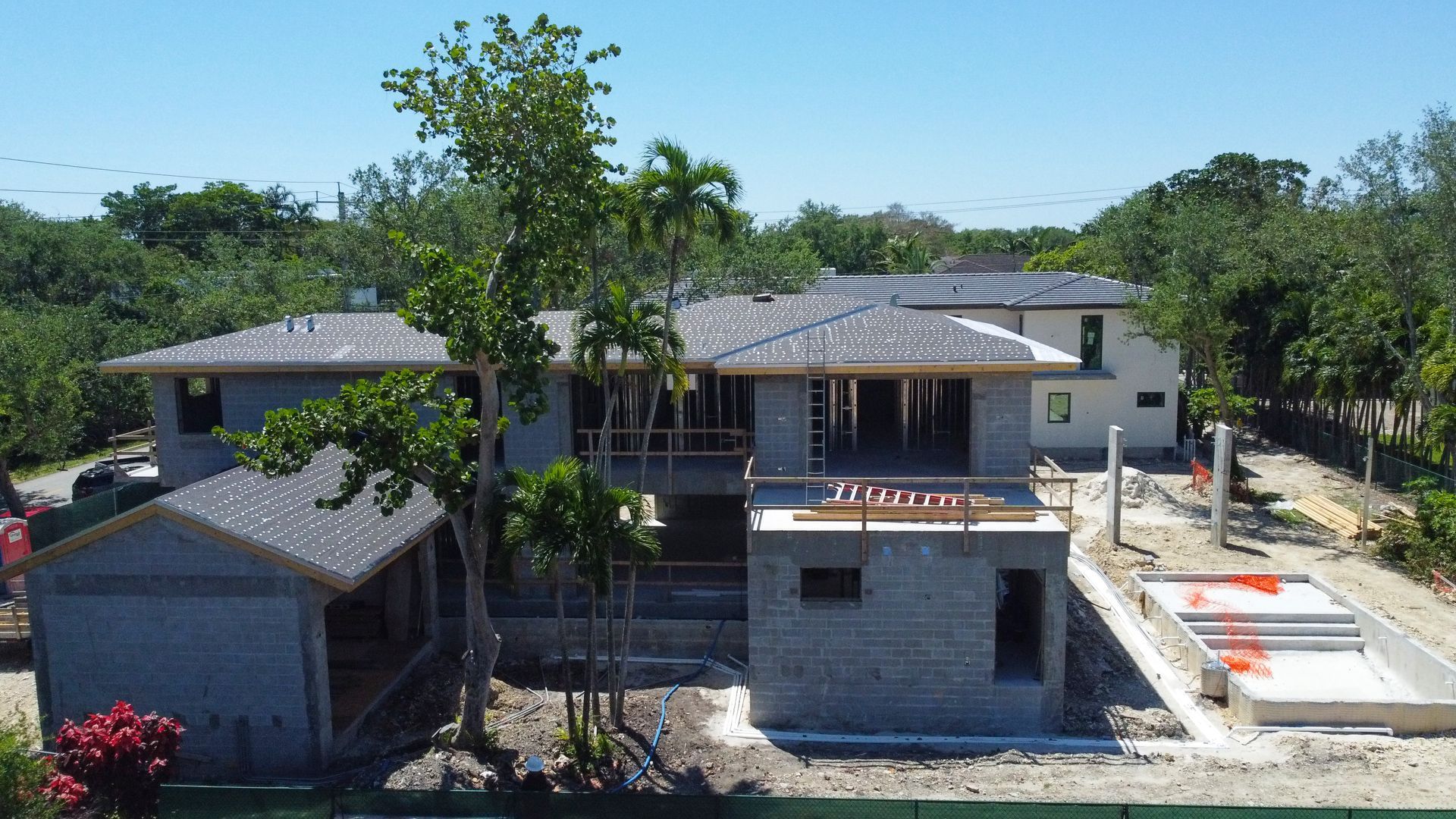 Construction site of a multi-level house with dark gray roof, surrounding trees, and a concrete pool.