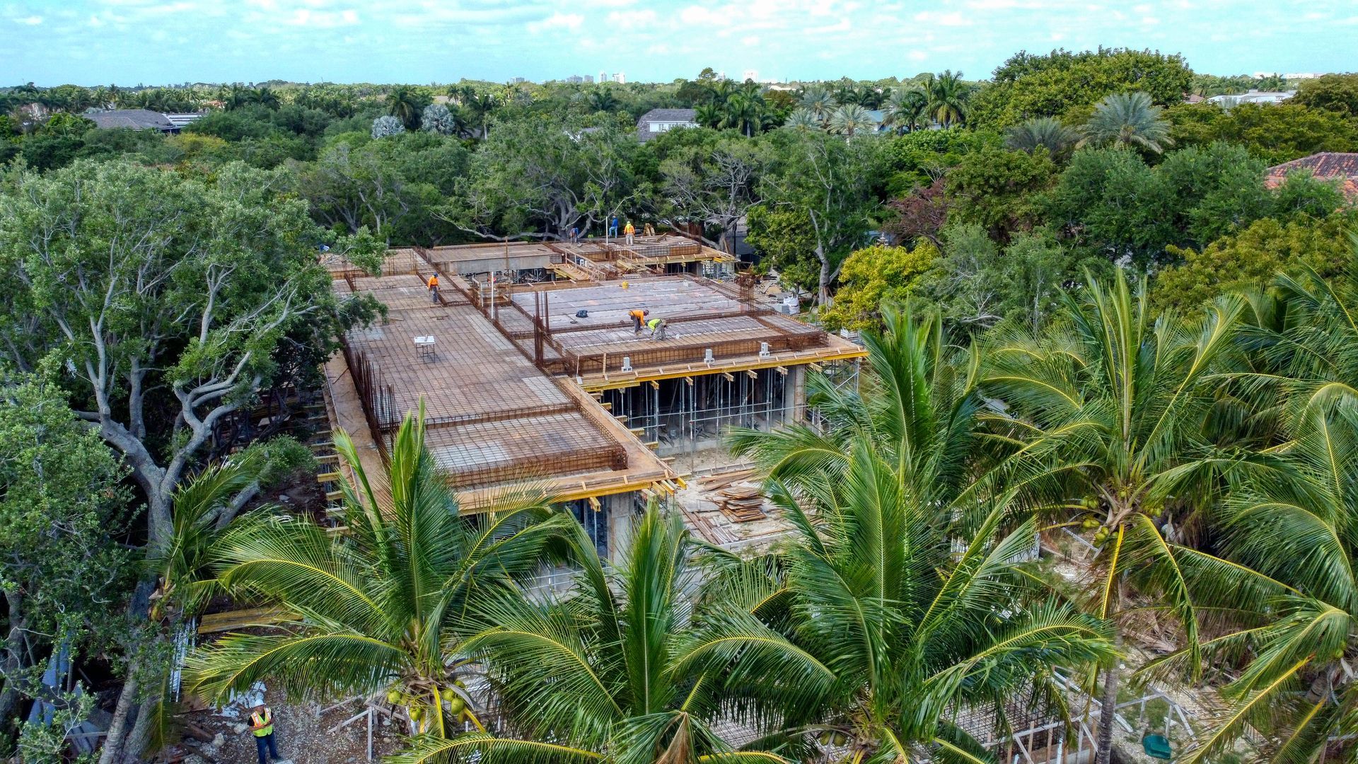 Aerial view of a building under construction, surrounded by lush green trees and palm fronds.