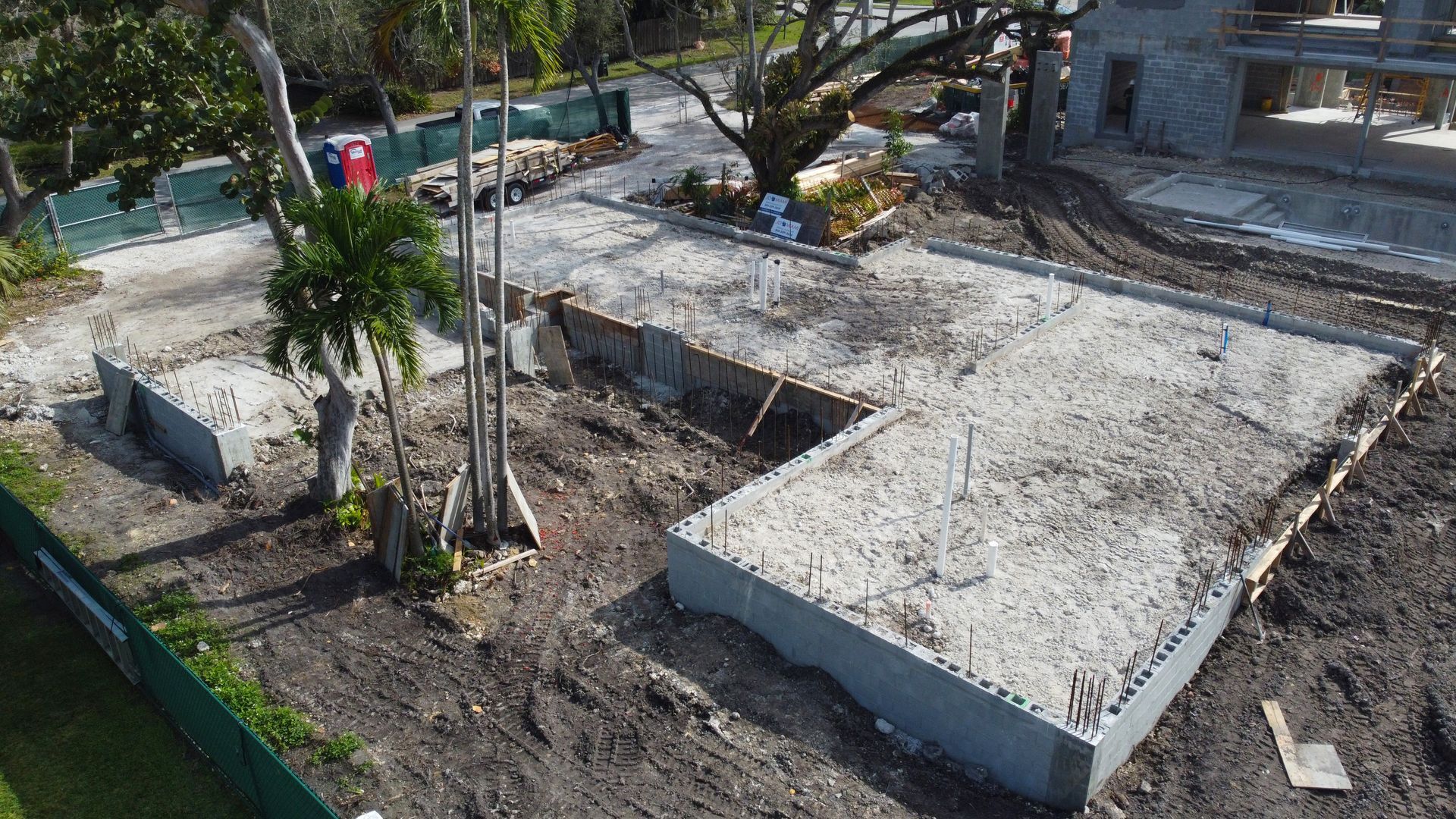Construction site with concrete foundations and dirt. A large tree and partially built building are visible.