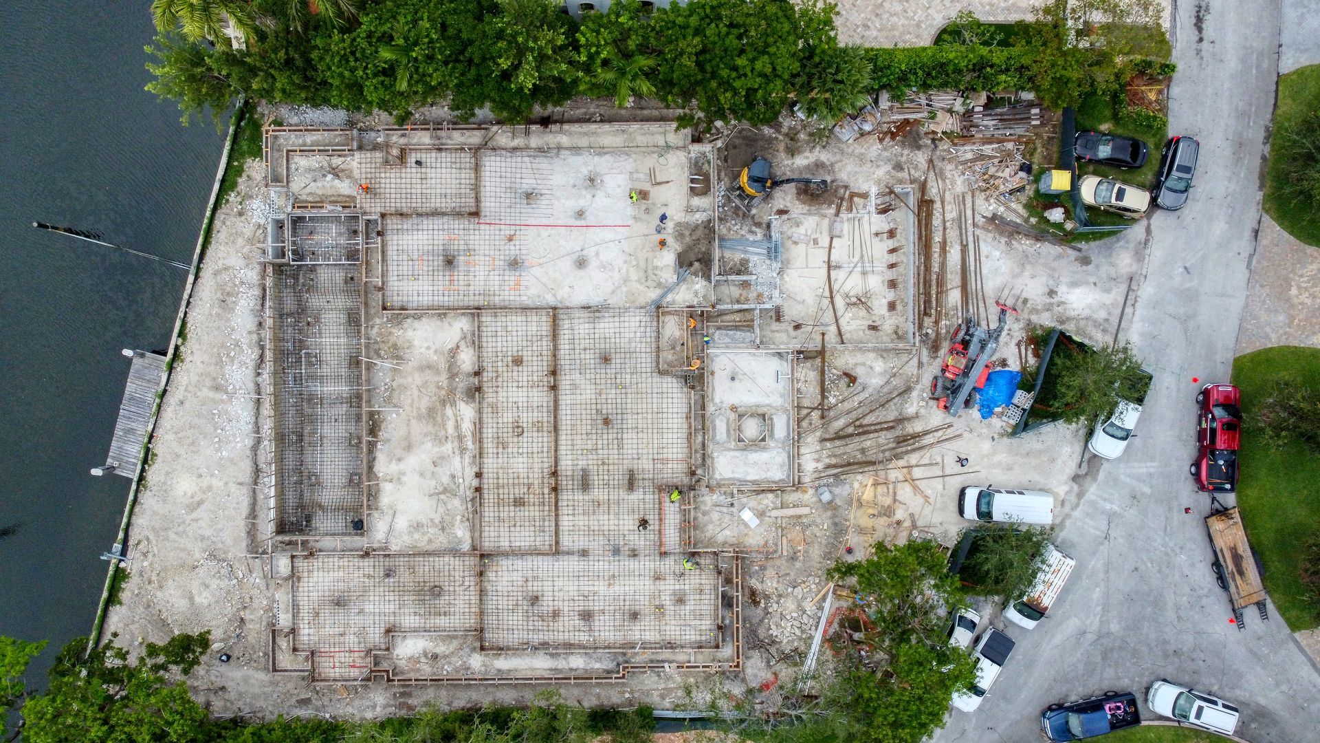 Aerial view of a construction site foundation; concrete slabs, surrounded by equipment and vehicles, near water.