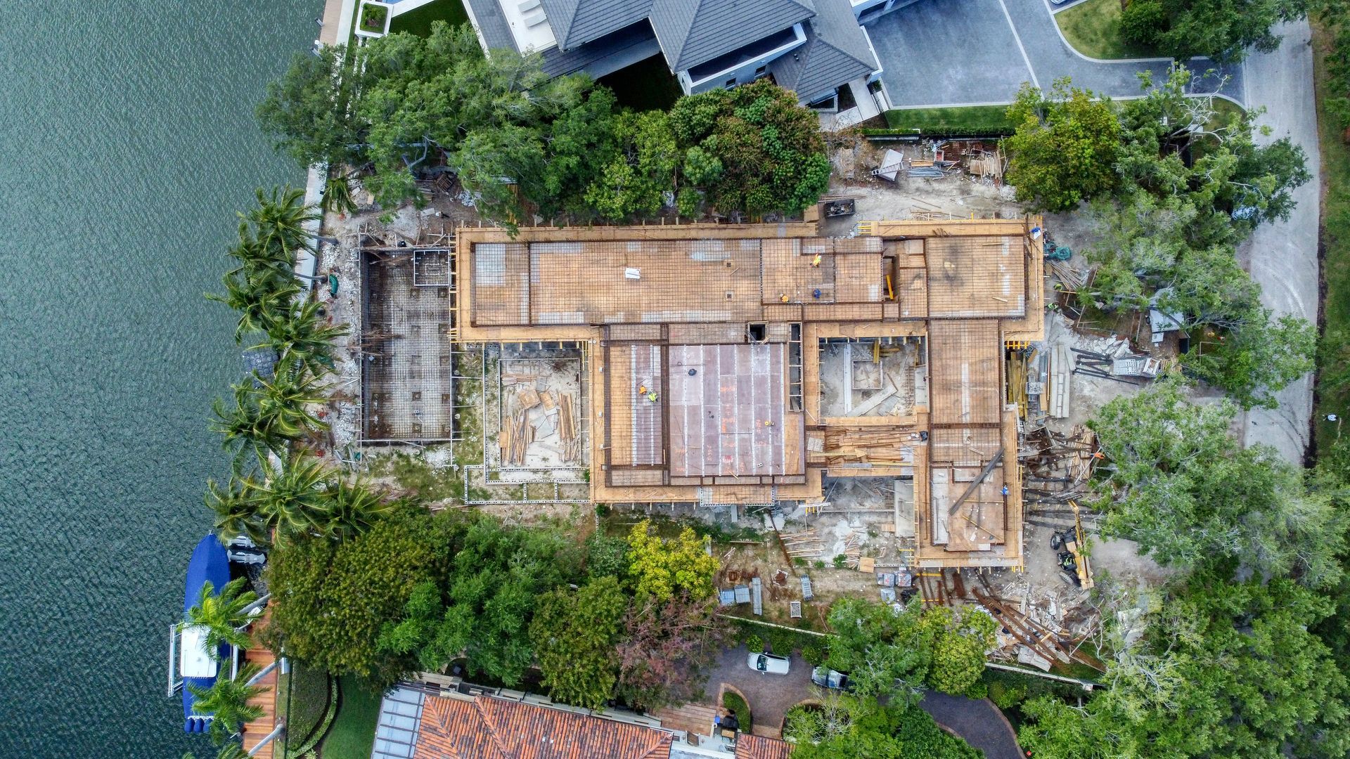 Aerial view of a house under construction; wooden frame on concrete foundation, surrounded by trees and water.