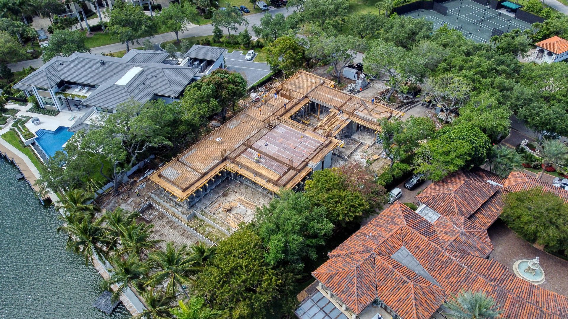 Aerial view of a home under construction with exposed wooden beams, surrounded by lush green trees and other houses.