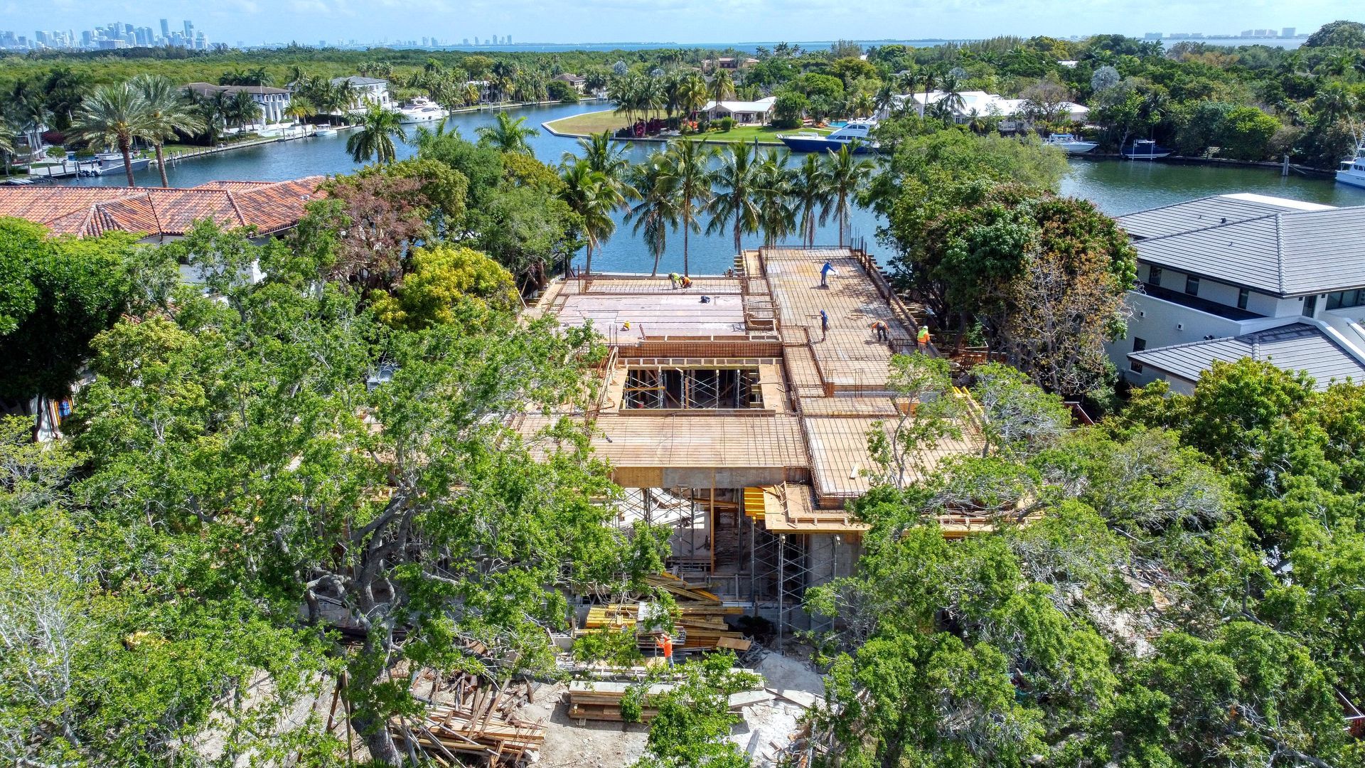 Construction site of a house with visible beams, surrounded by trees and waterfront homes.